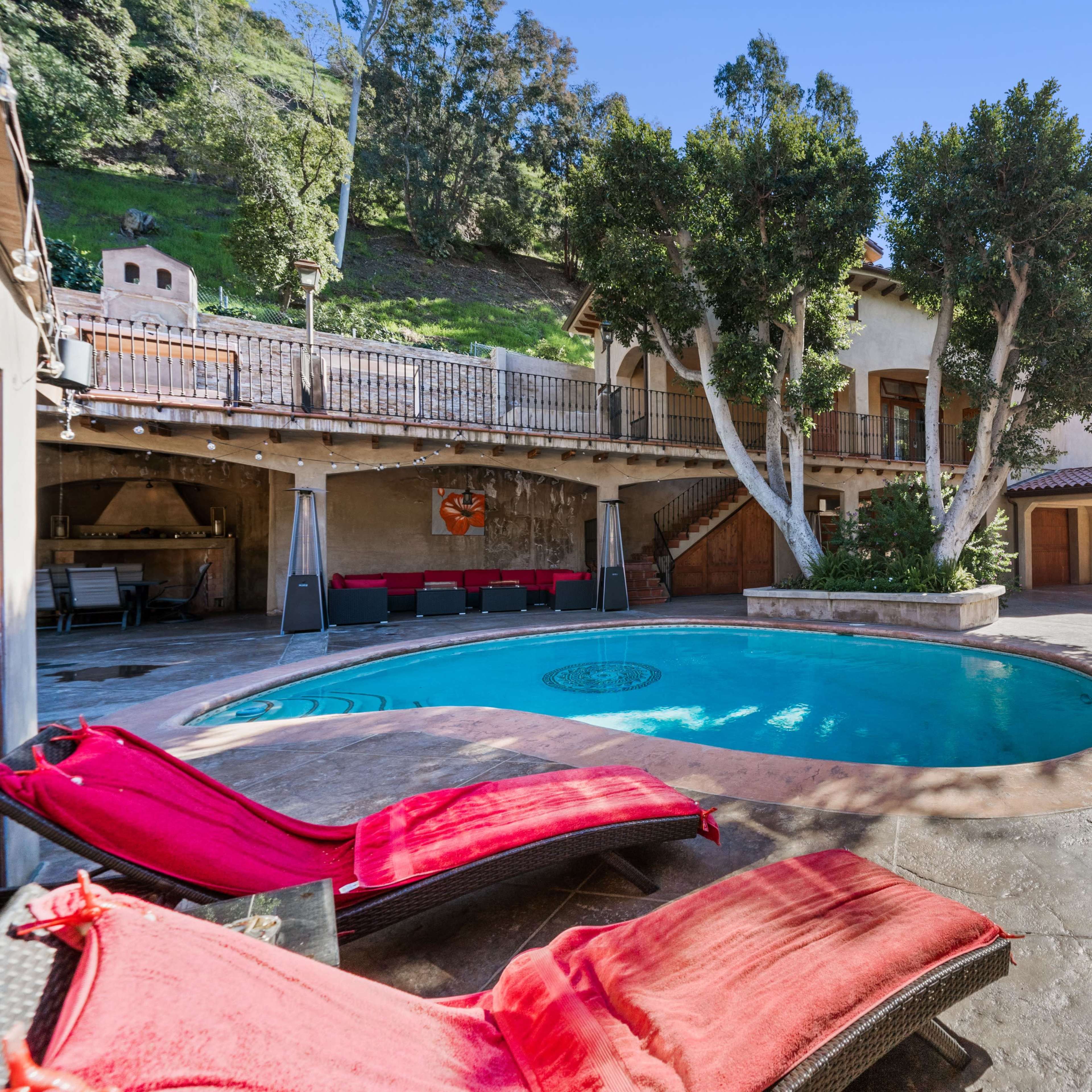 A residential backyard with a swimming pool surrounded by lounge chairs, a multi-level home, and a hillside in the background.