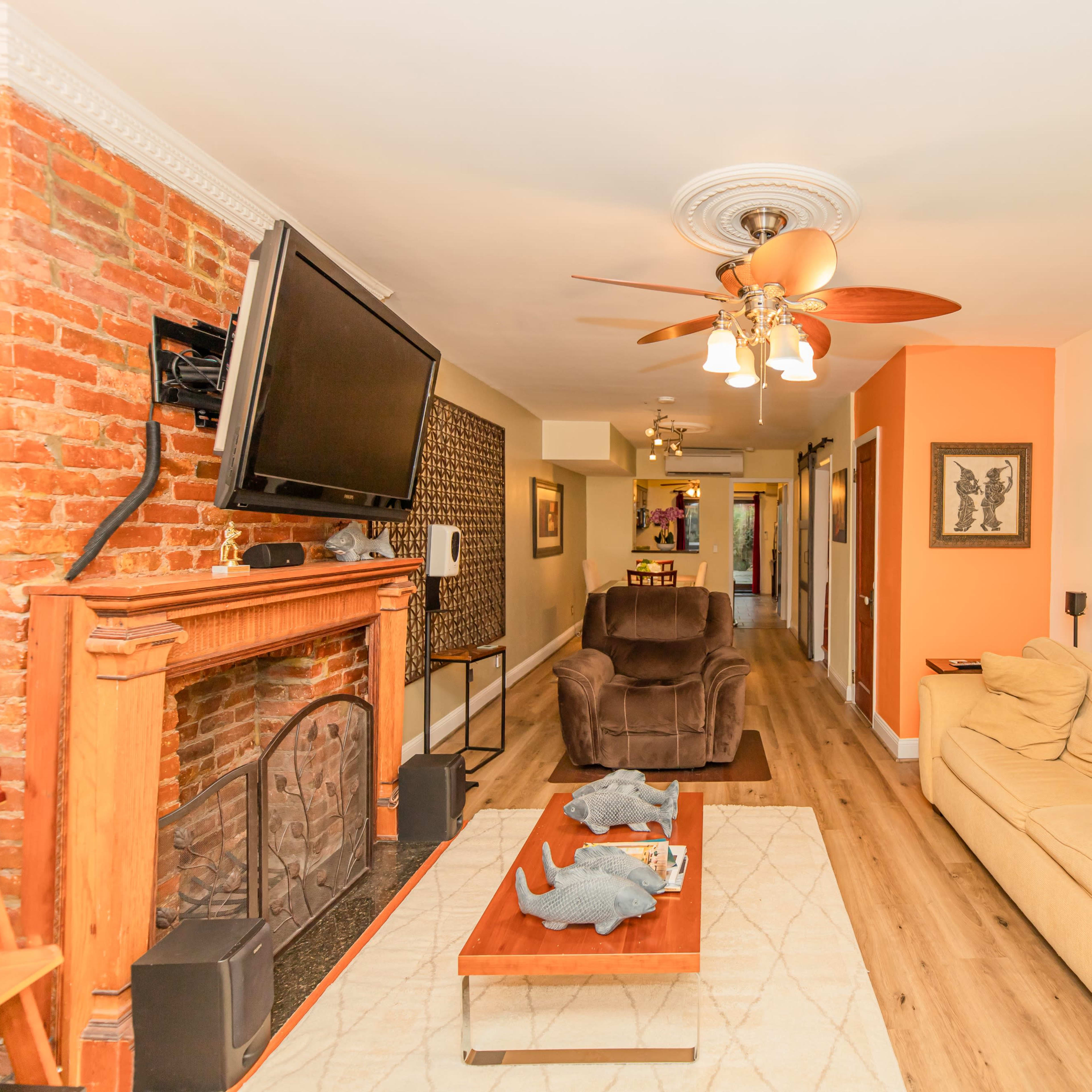 A living room with a brick fireplace, a flat-screen TV mounted on the wall, a beige sofa, and a wooden coffee table adorned with decorative items.