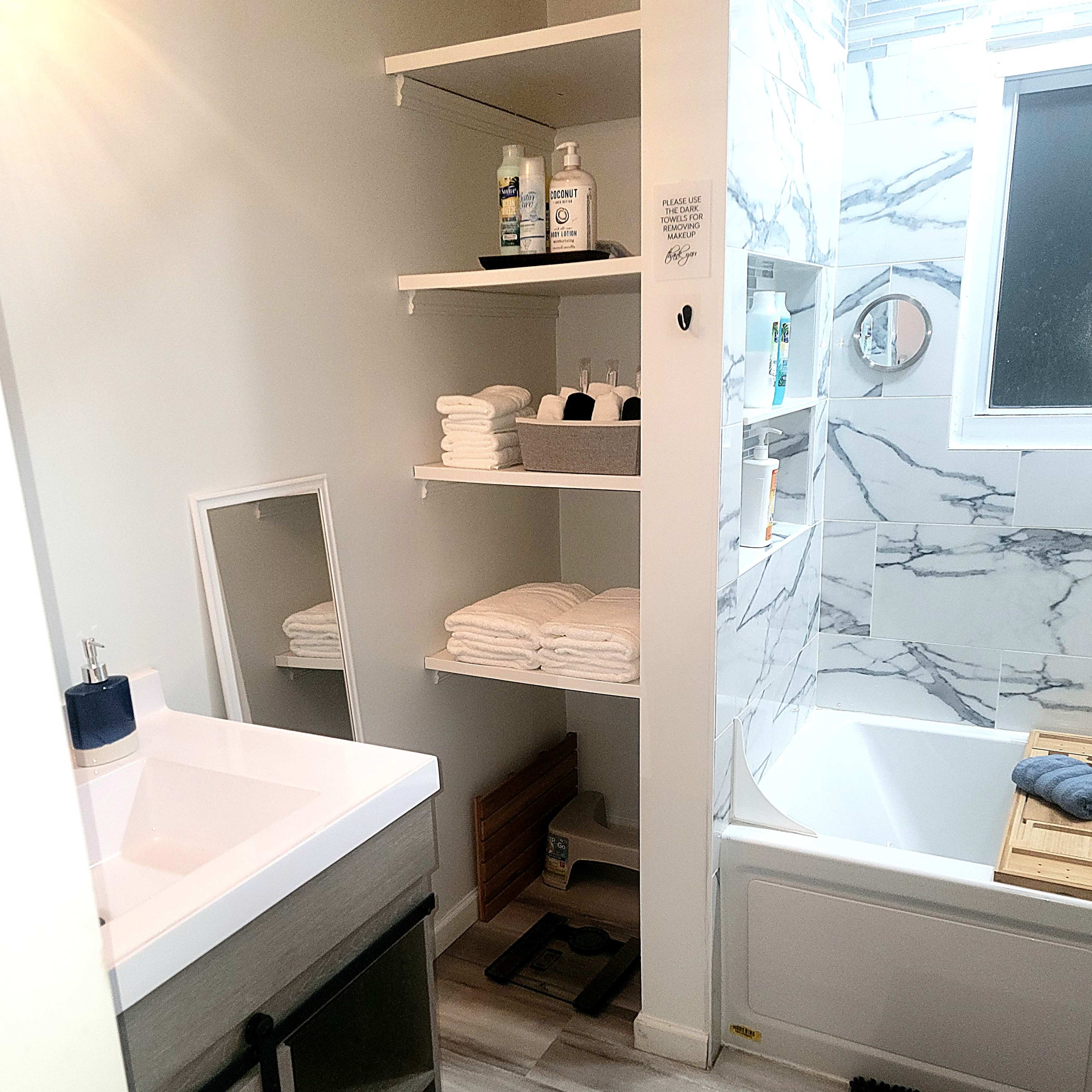 A modern bathroom featuring a bathtub with a shower, shelves with neatly stacked towels, a sink with a mirror, and a gray cabinet.