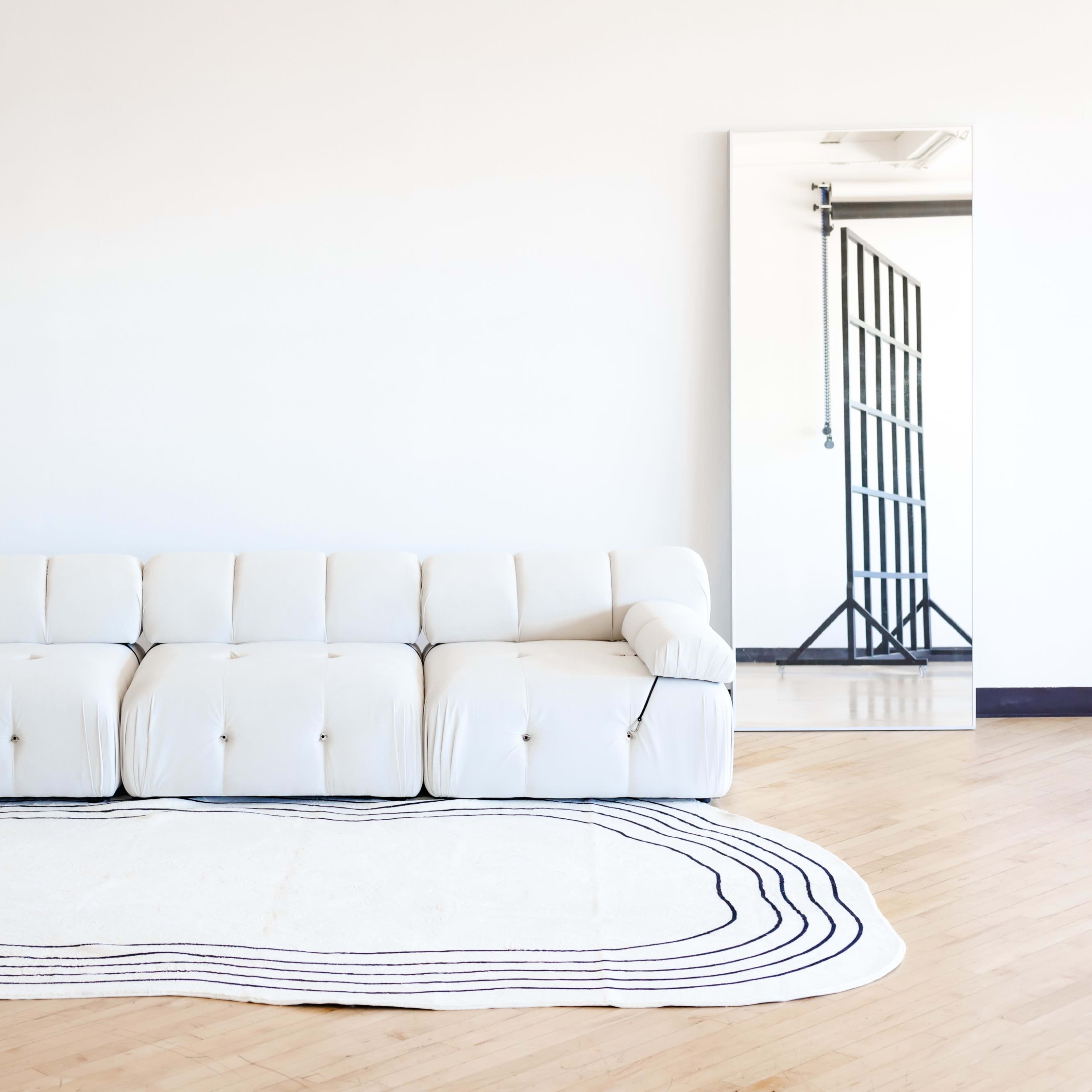 A white sofa and an orange chair are positioned in a minimalistic room with a large mirror on the wall.