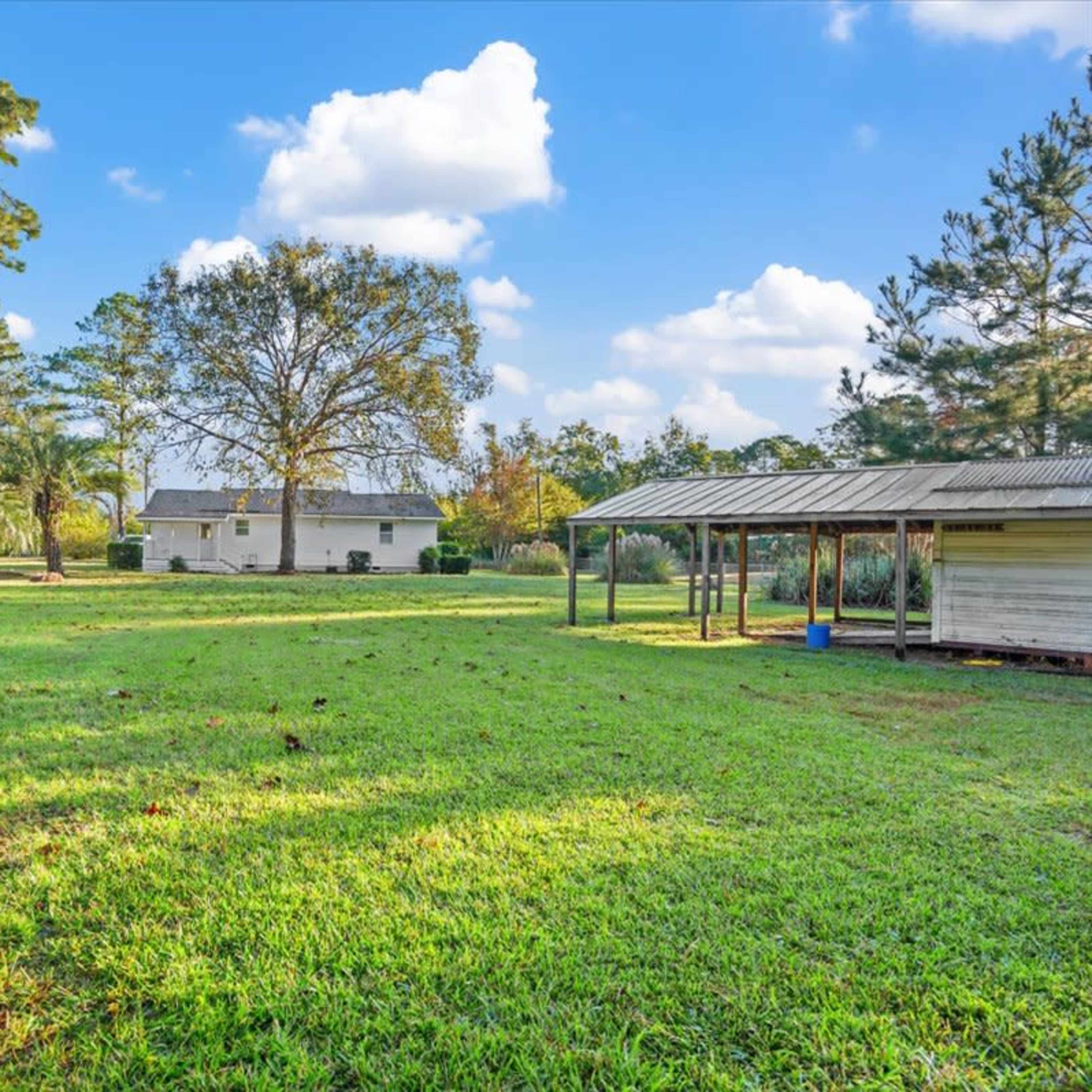 A spacious yard with a house in the background, surrounded by trees and a covered structure on one side.