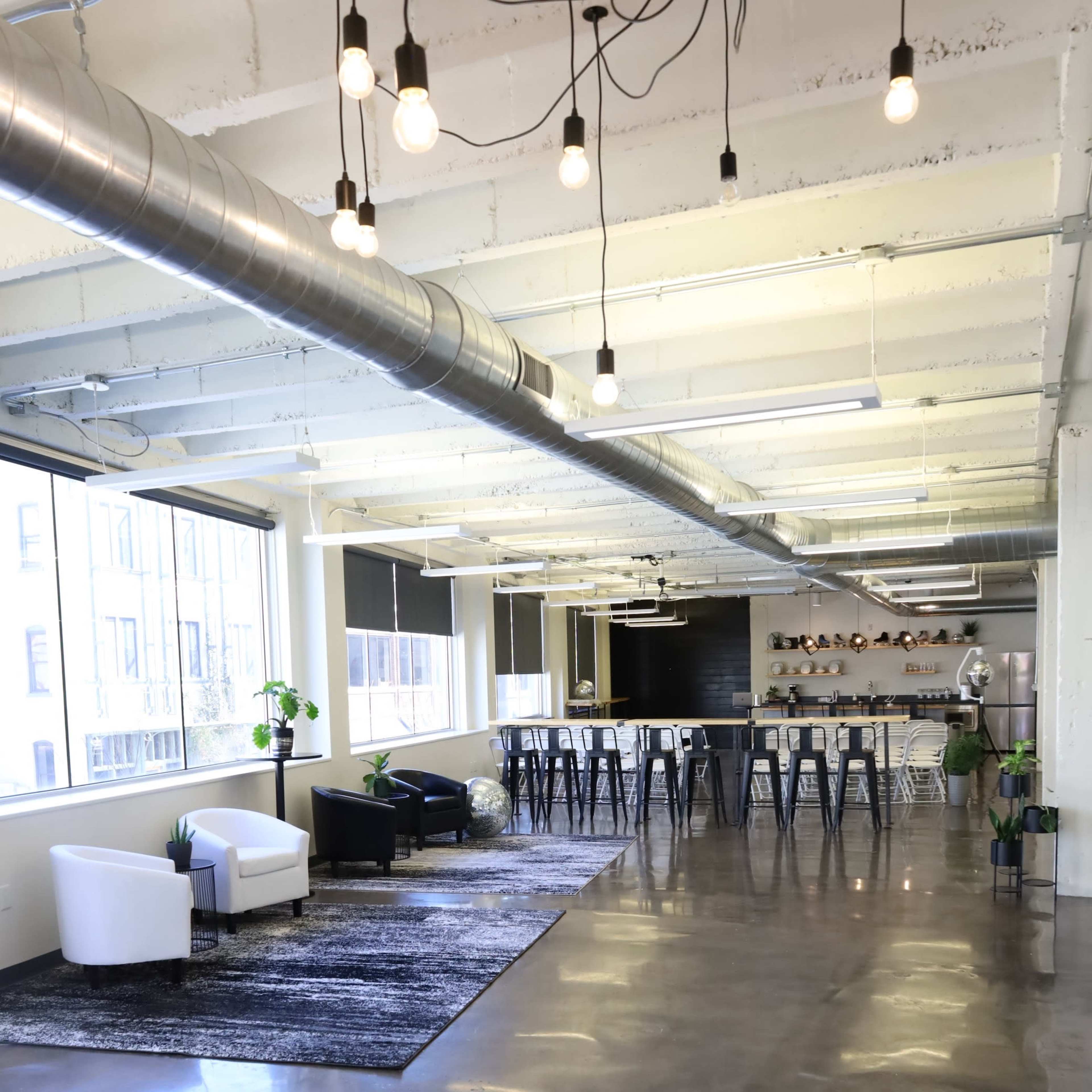 A spacious, modern office interior with polished concrete floors, a seating area with two armchairs, and a long bar-style table with metal stools.