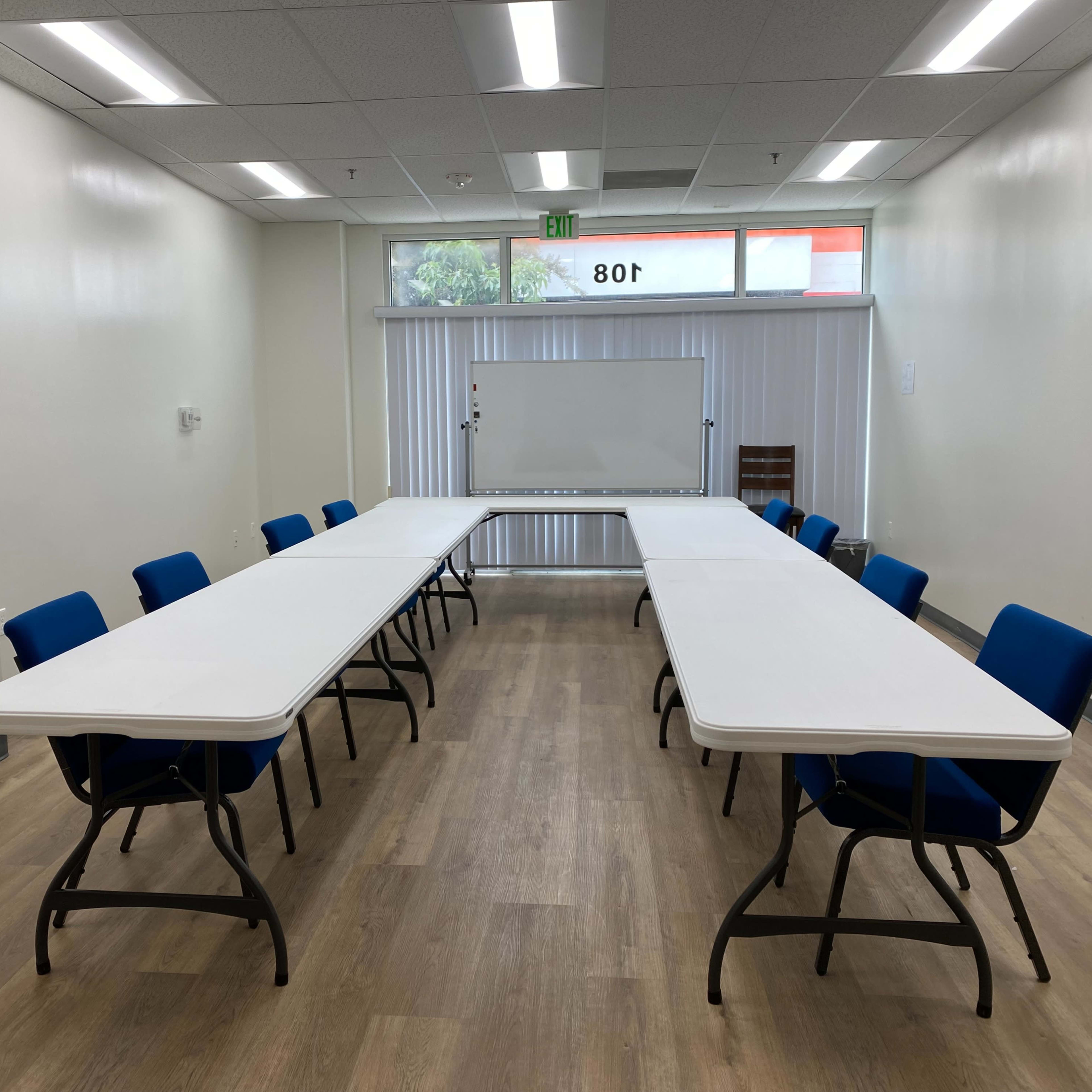 A training room features two sets of rectangular tables arranged in a U-shape, surrounded by blue chairs, with large windows allowing natural light.