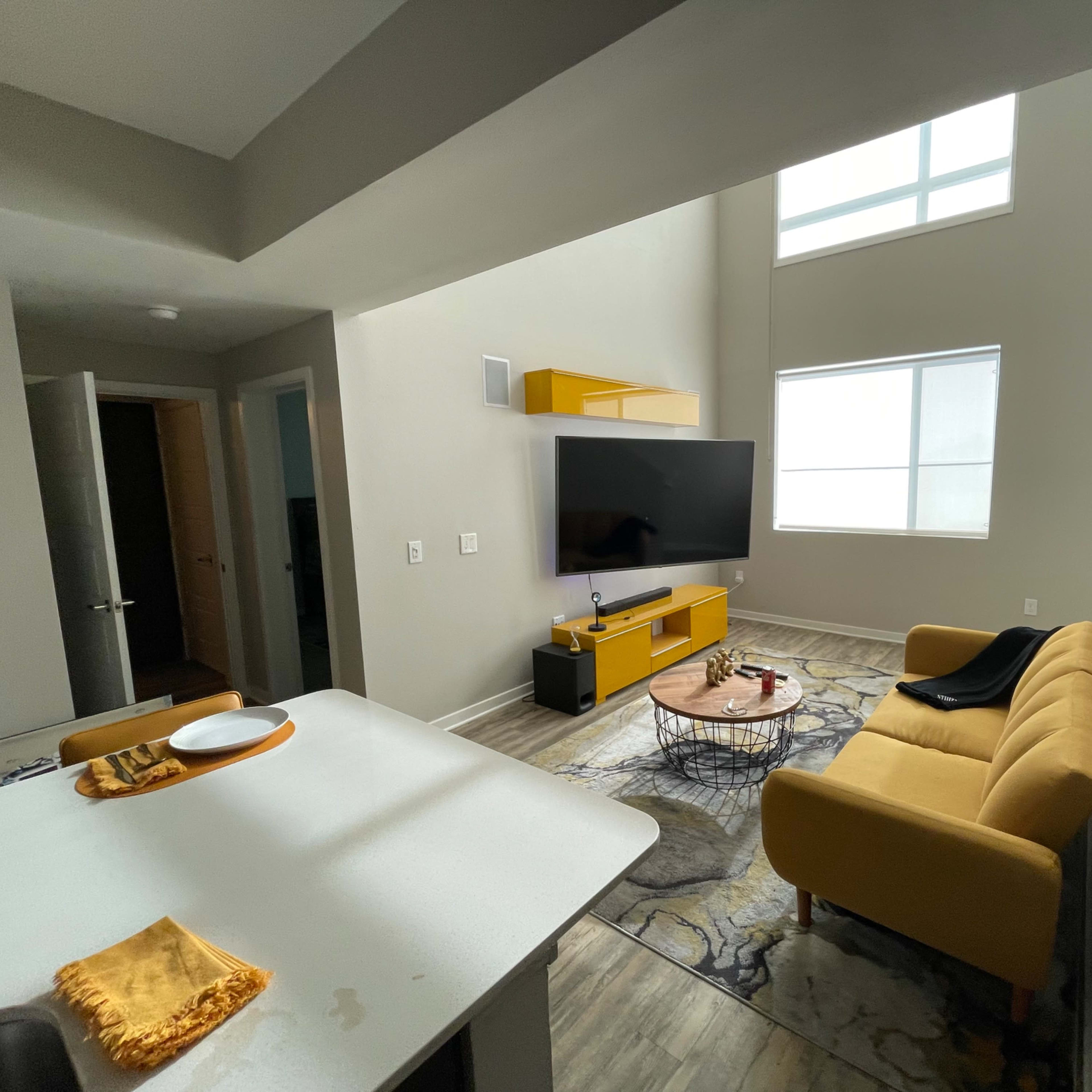A modern living room with a yellow sofa, a wall-mounted television, and a large window allowing natural light to enter.