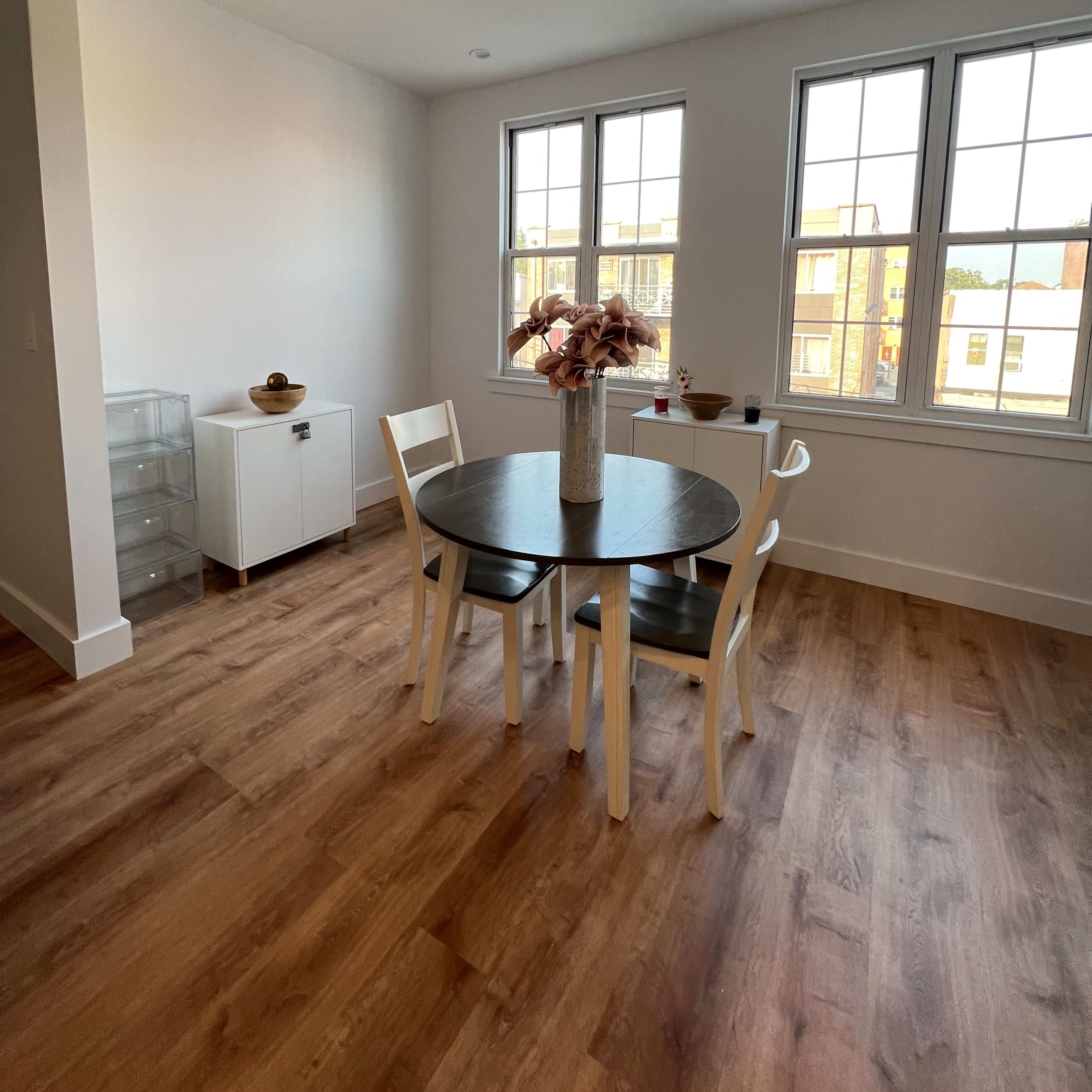 A dining area features a round table with two chairs, a floral centerpiece, and large windows allowing natural light to enter.