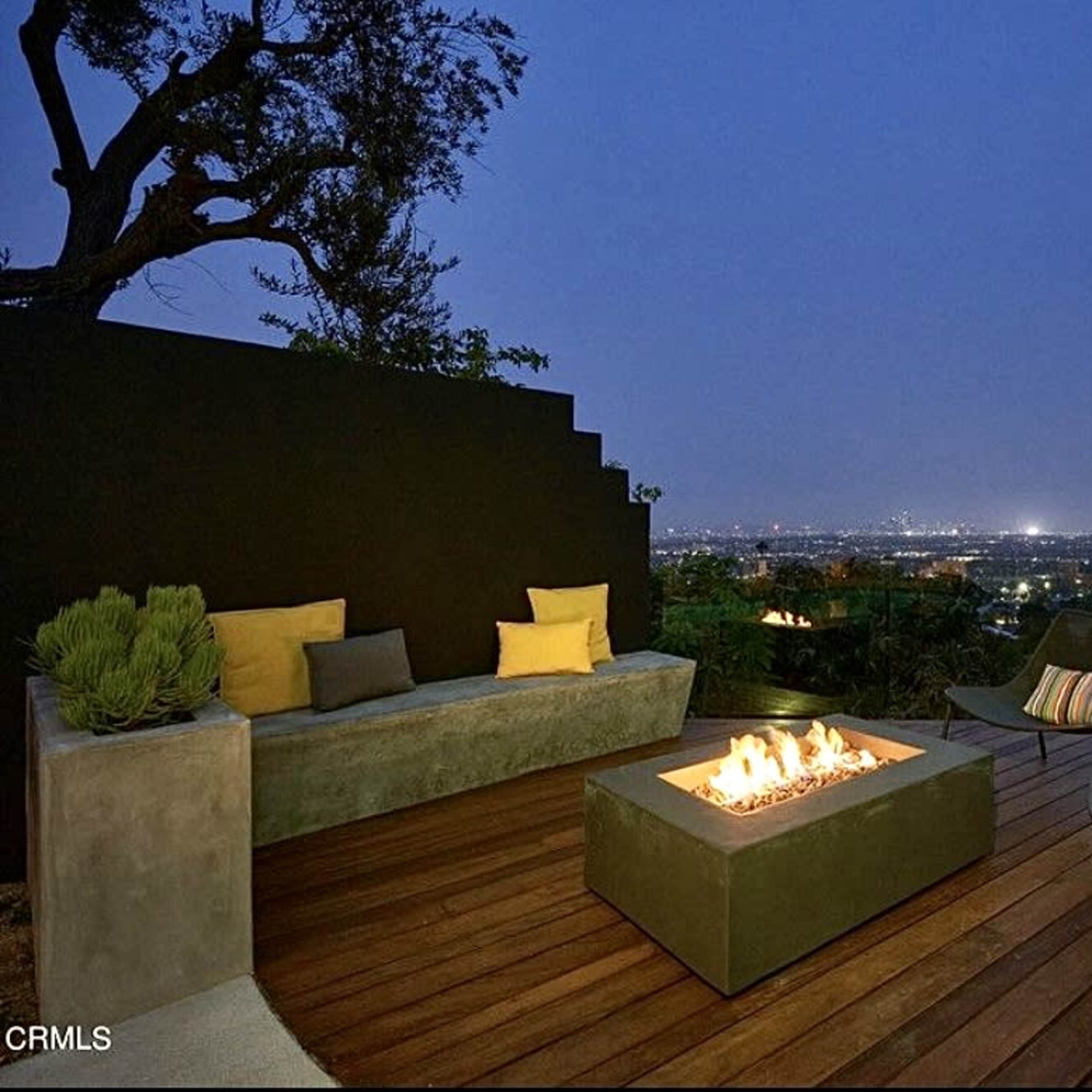 A modern outdoor seating area features a fire pit surrounded by comfortable chairs, with a panoramic view of a city skyline at dusk.