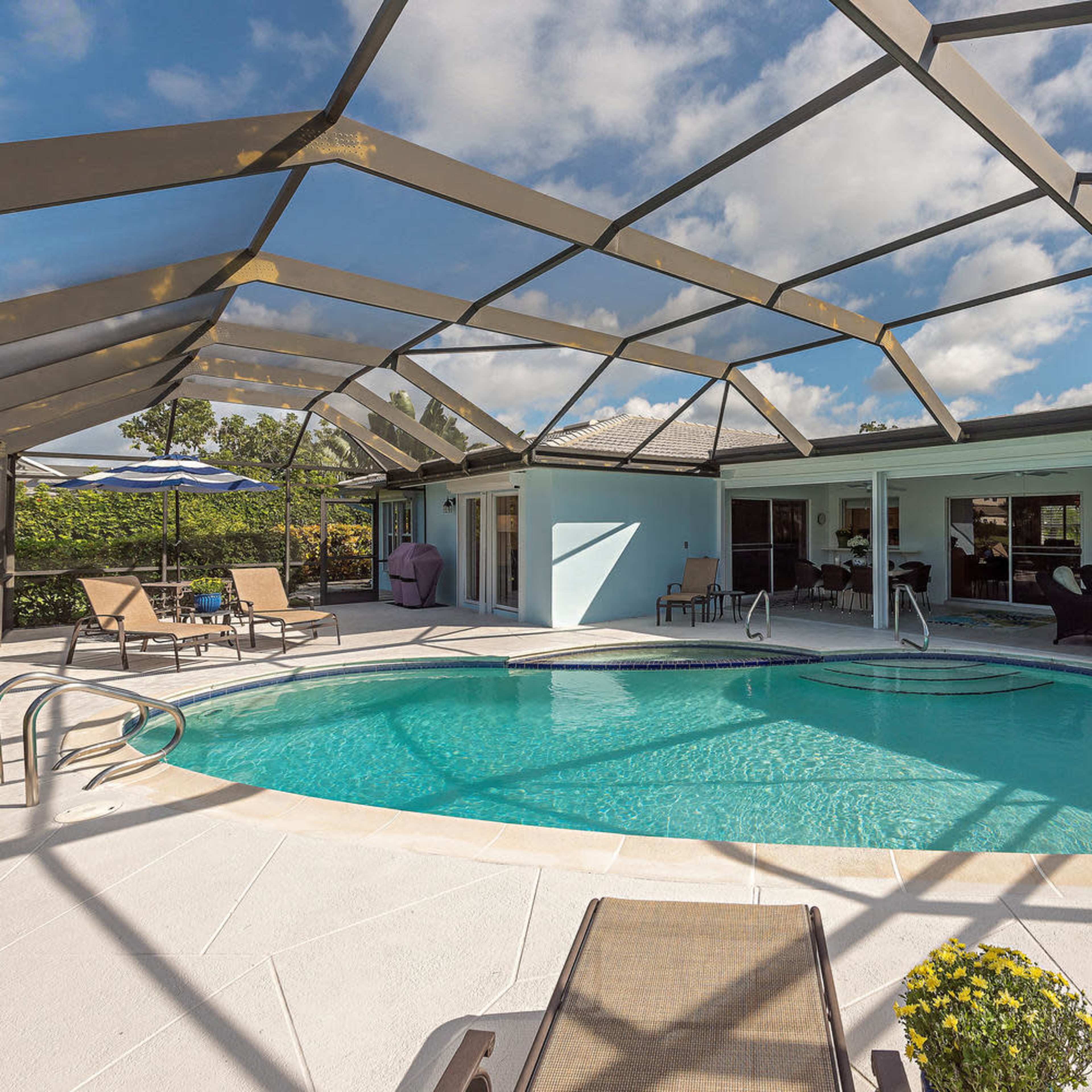 A screened-in swimming pool surrounded by lounge chairs and a patio area with a view of a house and landscaped greenery.