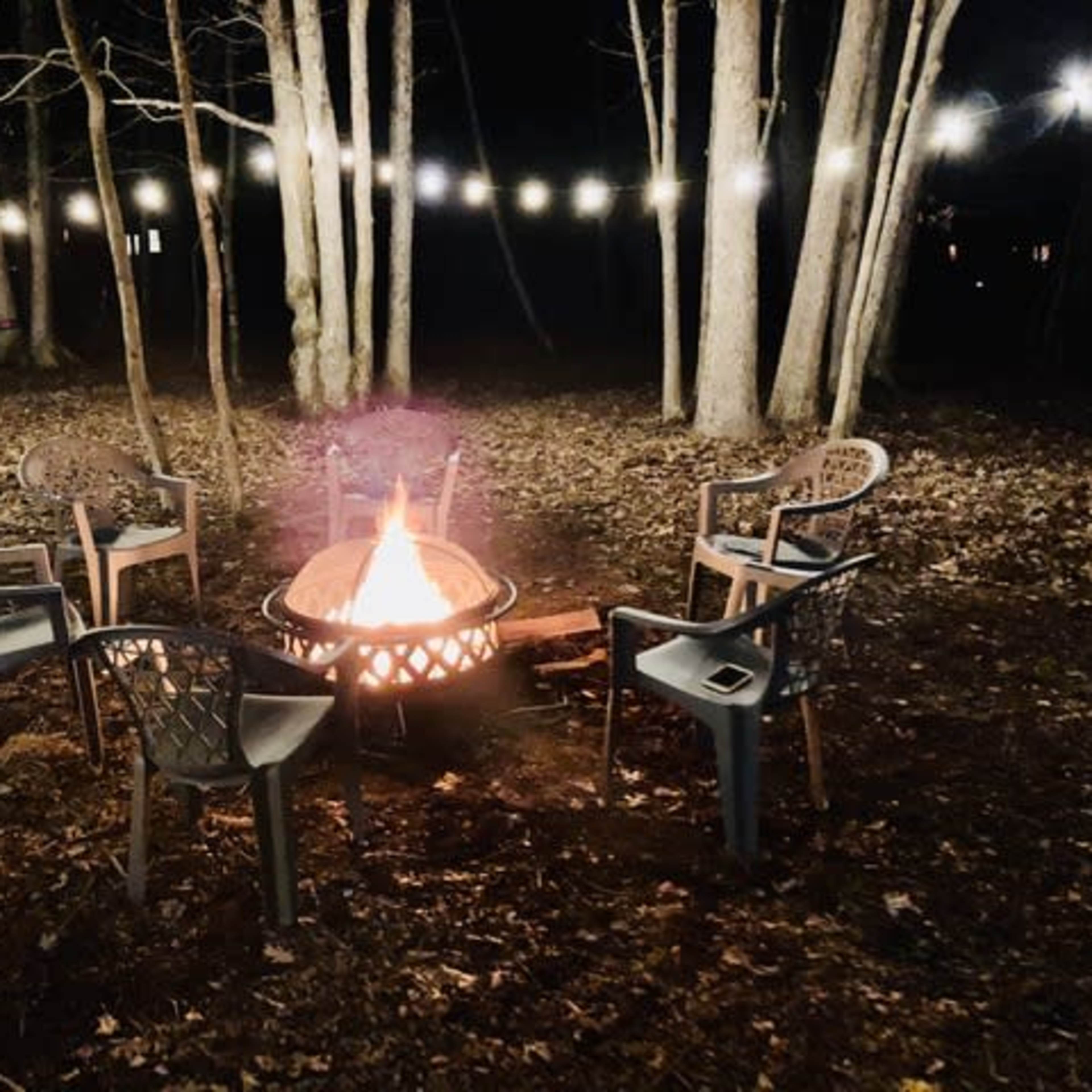 A fire pit surrounded by five chairs sits in a wooded area at night, illuminated by string lights above.