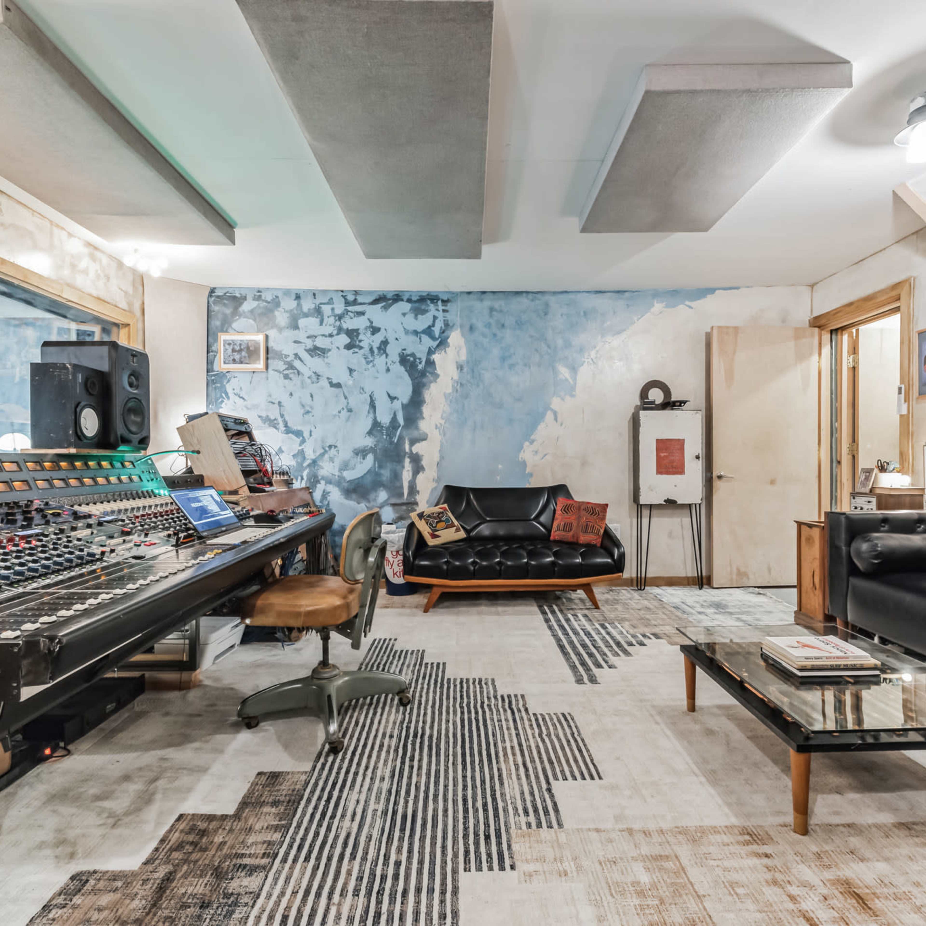 A sound studio features a mixing console, two couches, and a patterned rug against a textured wall.