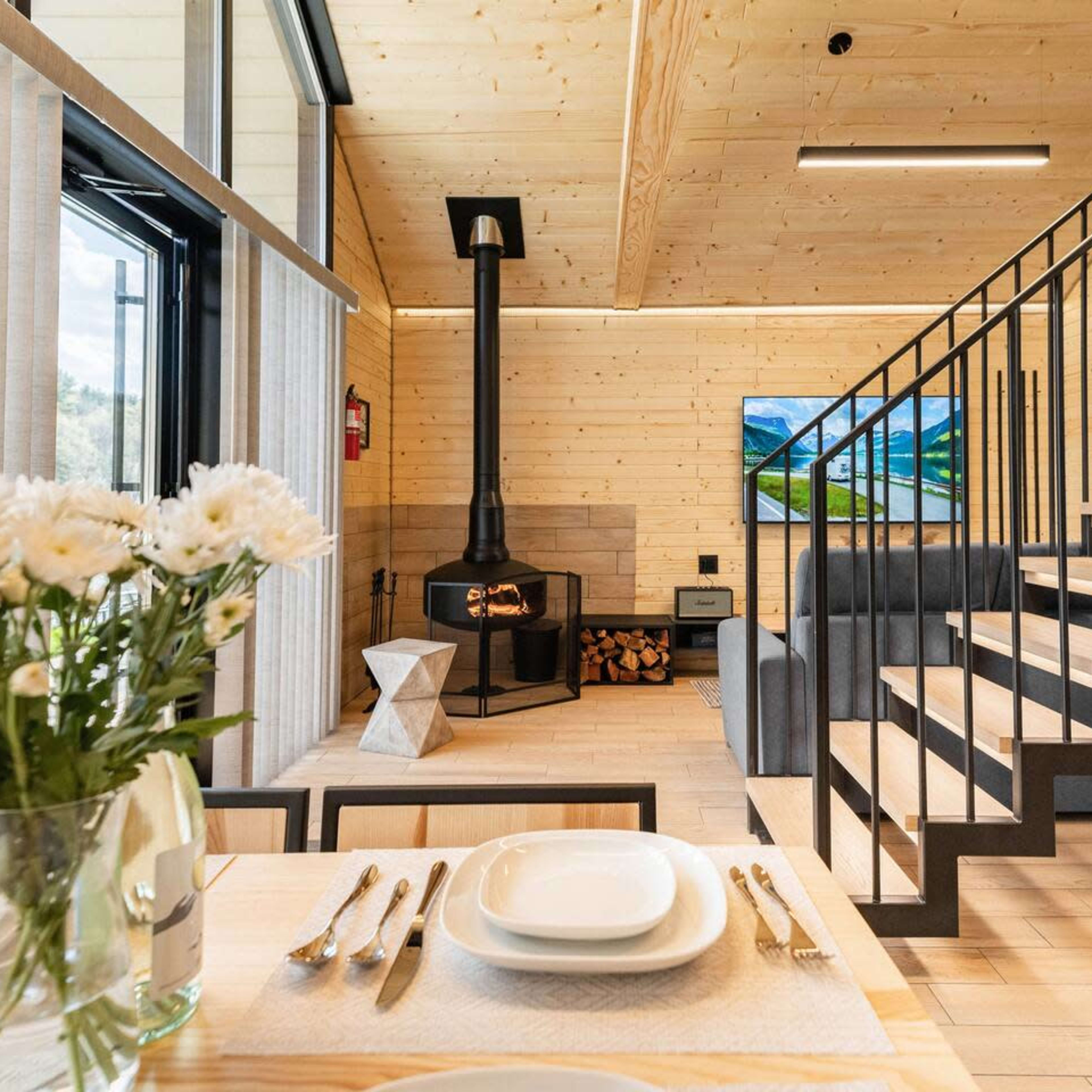 A modern, wooden interior living space featuring a staircase, a fireplace, a dining table with flowers, and large windows.