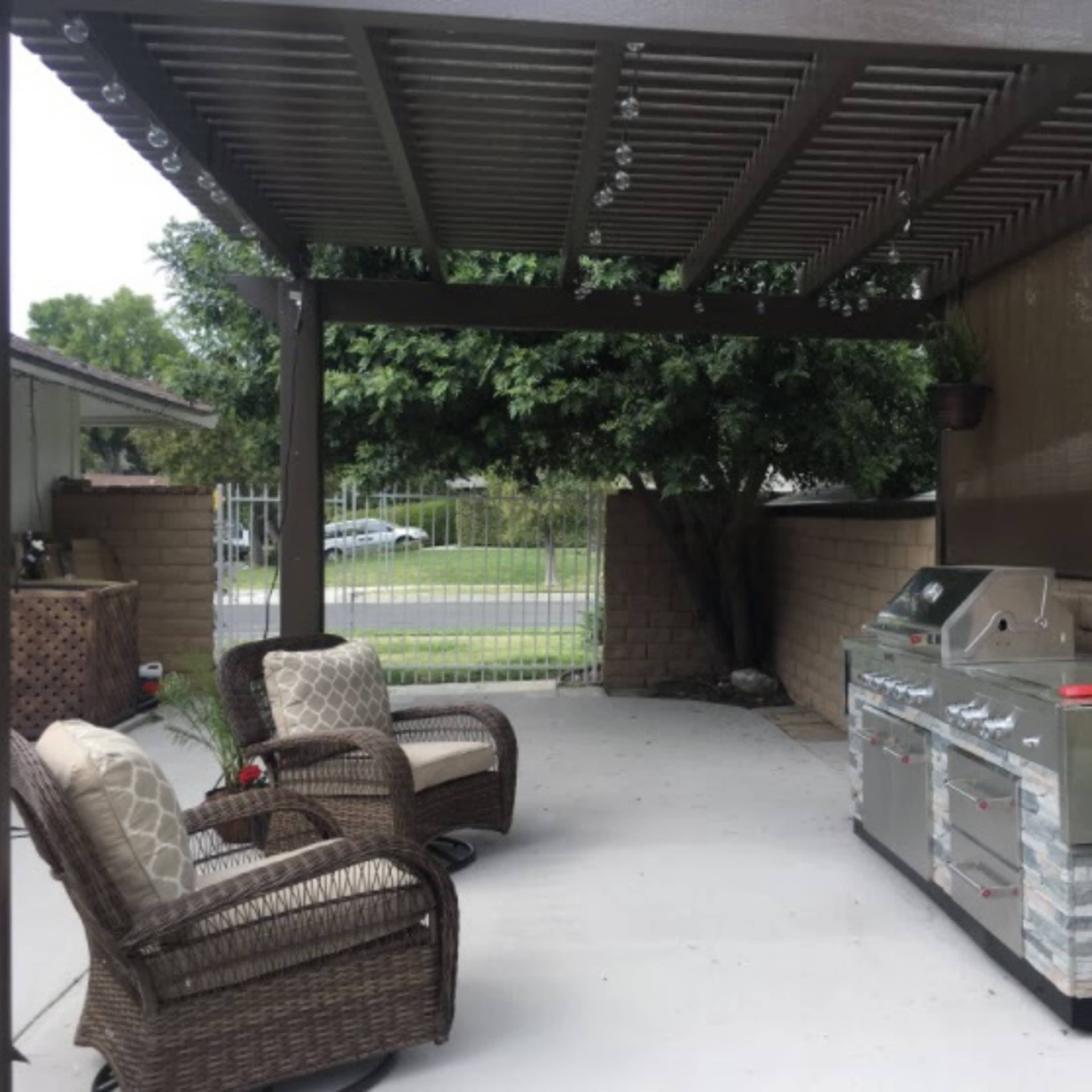 A shaded outdoor patio with two wicker chairs and a stainless steel barbecue grill against a stone-accented wall.