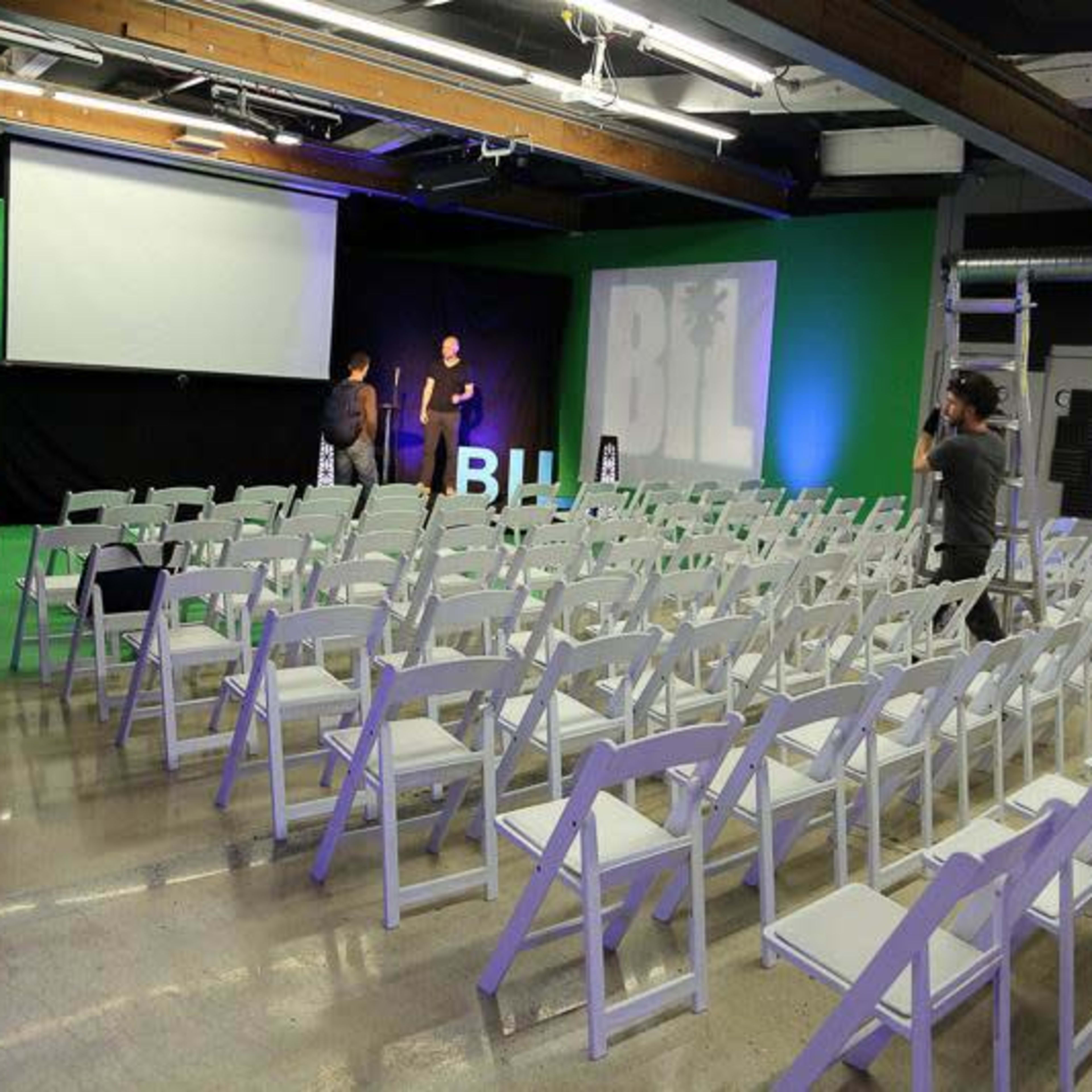 A room with rows of white chairs arranged for an event, featuring a projector screen and green backdrop.