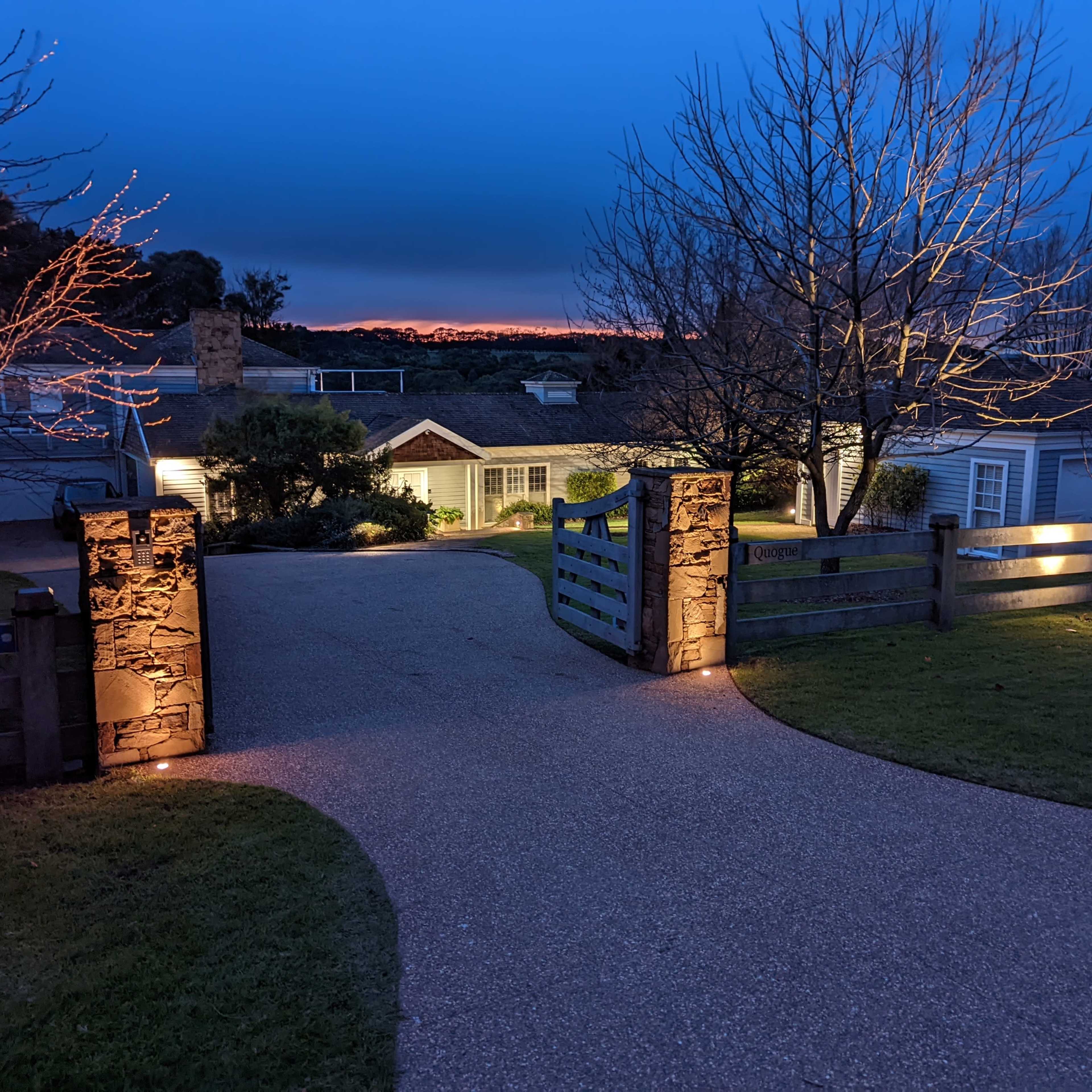 A well-lit driveway leads to a house at dusk, framed by stone pillars and trees.