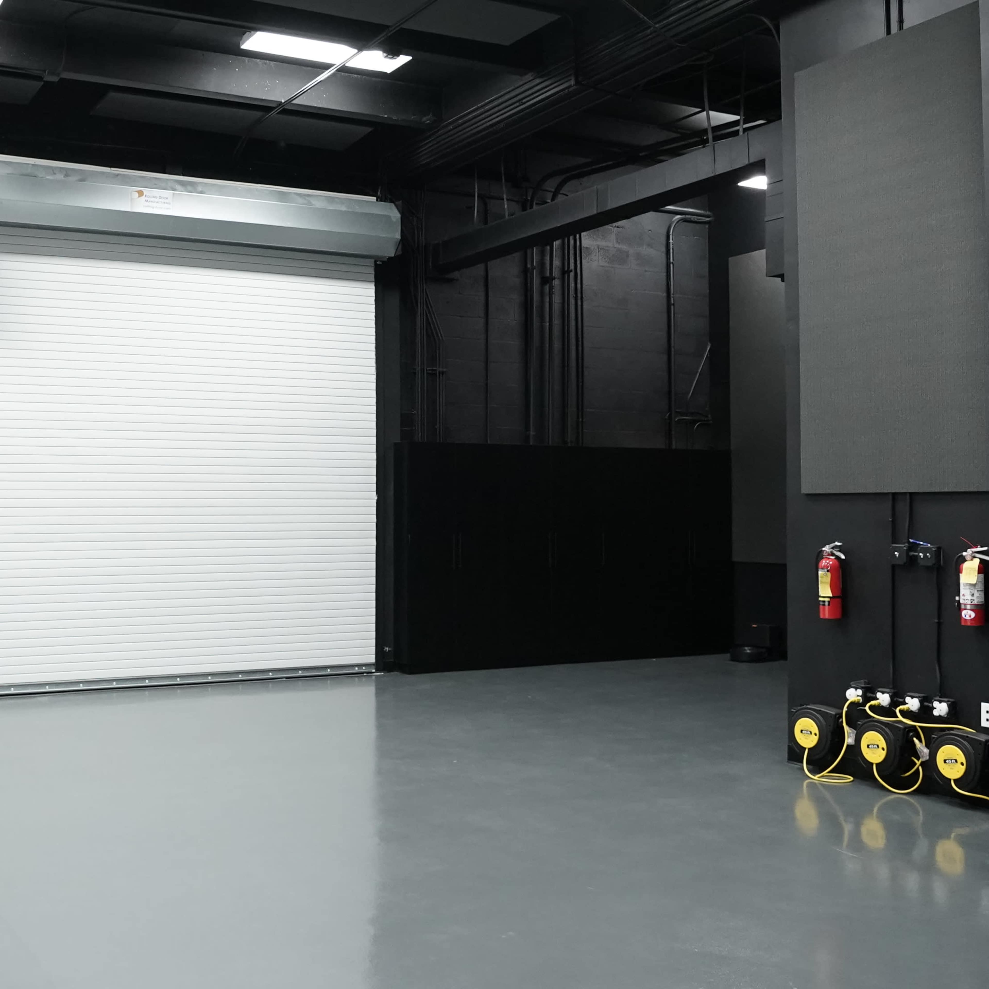 A large, empty industrial space with a closed rolling garage door and minimal equipment against black walls.