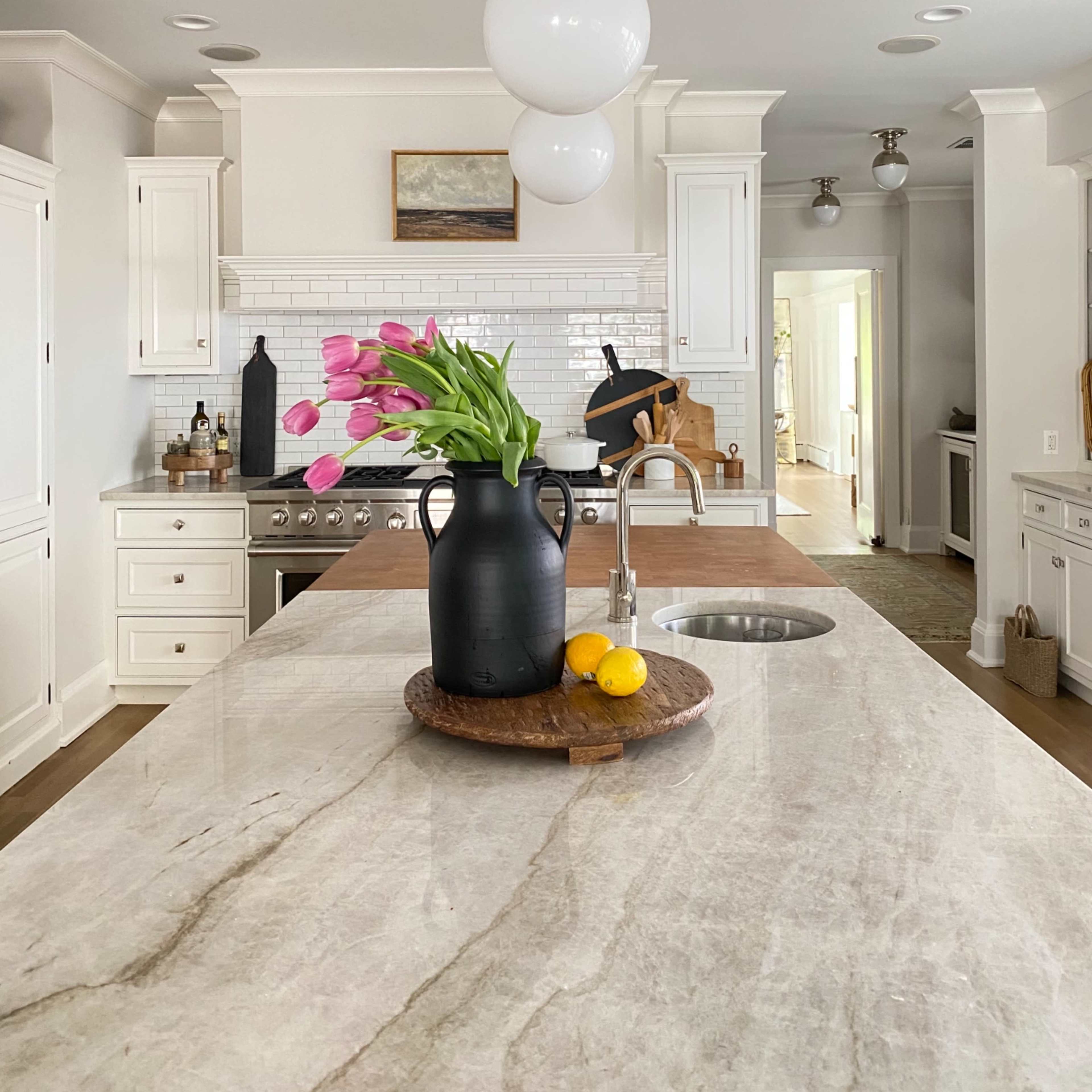 A modern kitchen features a large island with a wooden tray holding a black vase of pink tulips and yellow lemons, surrounded by white cabinetry and stainless steel appliances.
