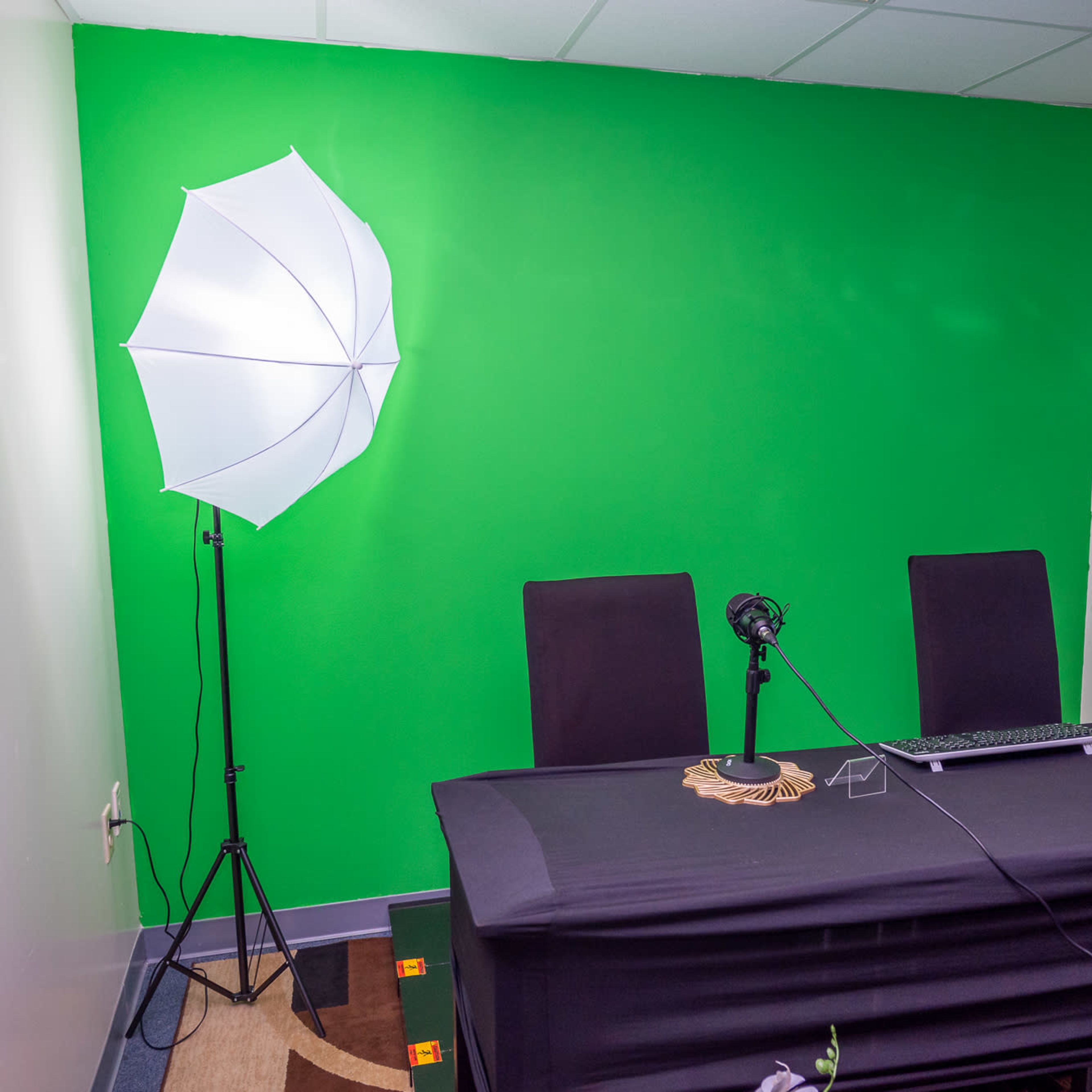 A small studio with a green screen wall, two chairs at a table, microphones on stands, and a monitor displaying a sports game.