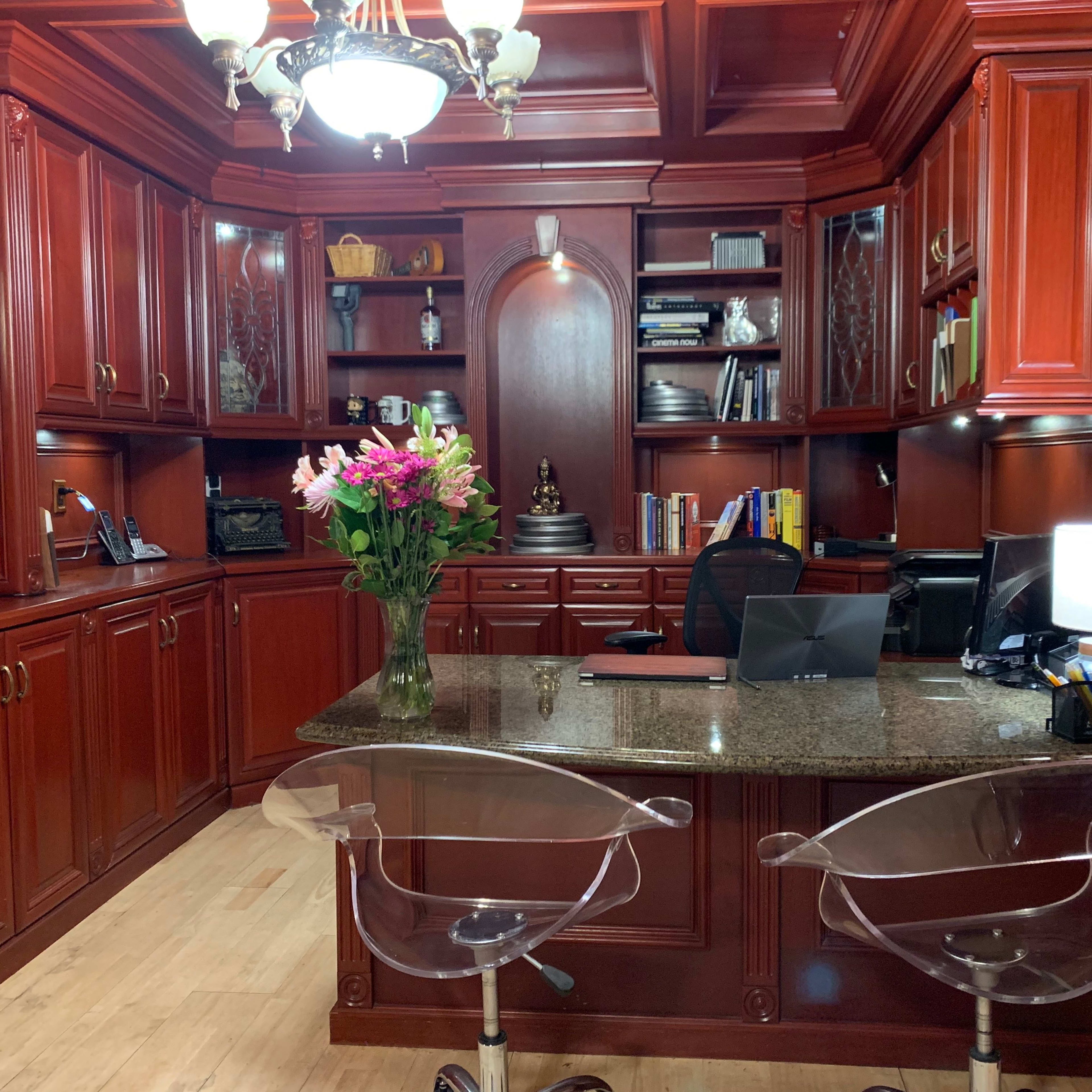 A wood-paneled office with a granite countertop, clear acrylic chairs, and shelves filled with books and decorative items, illuminated by a chandelier.