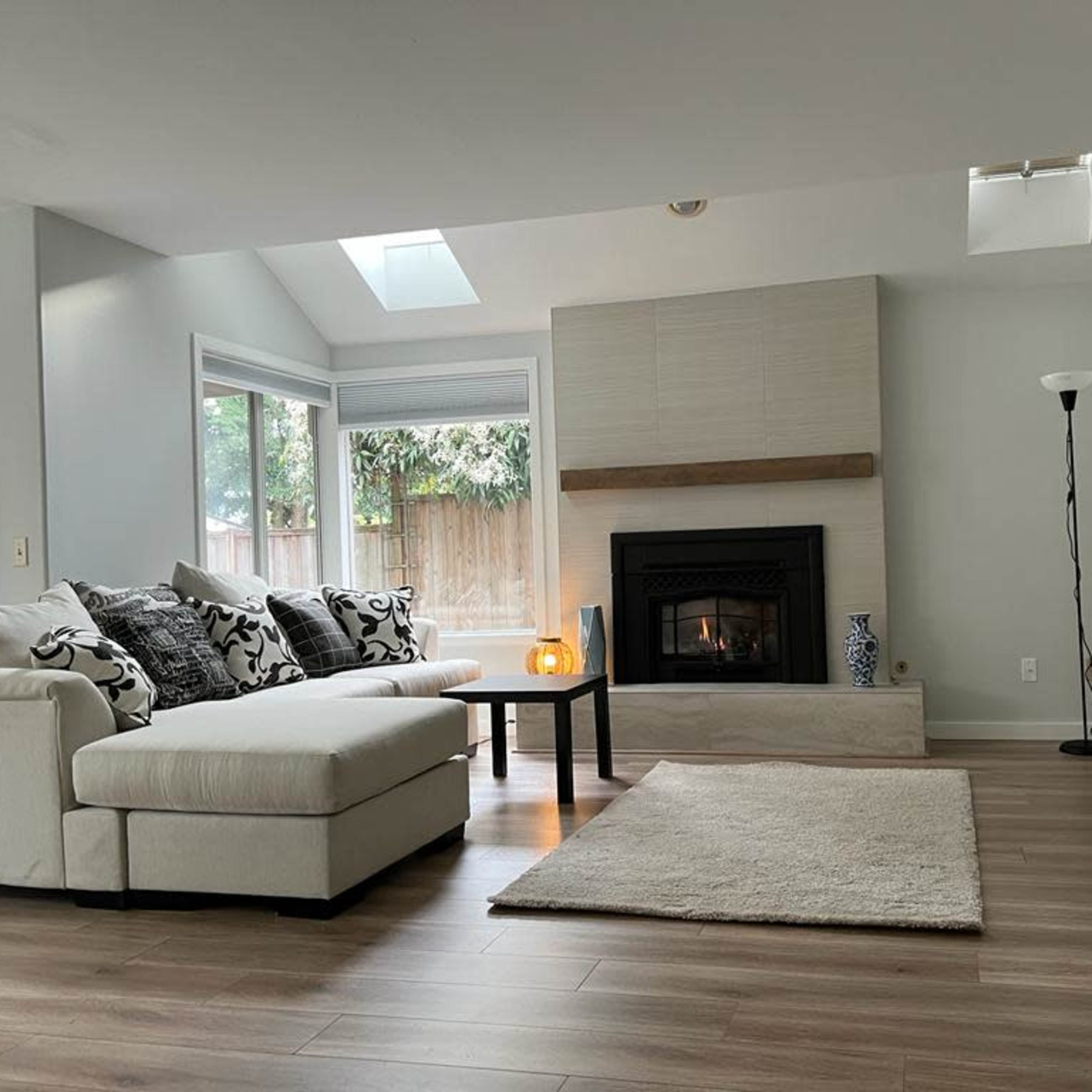 A modern living room features a white sectional sofa, a coffee table, a fireplace, and a television on a wooden console, with large windows allowing natural light to enter.