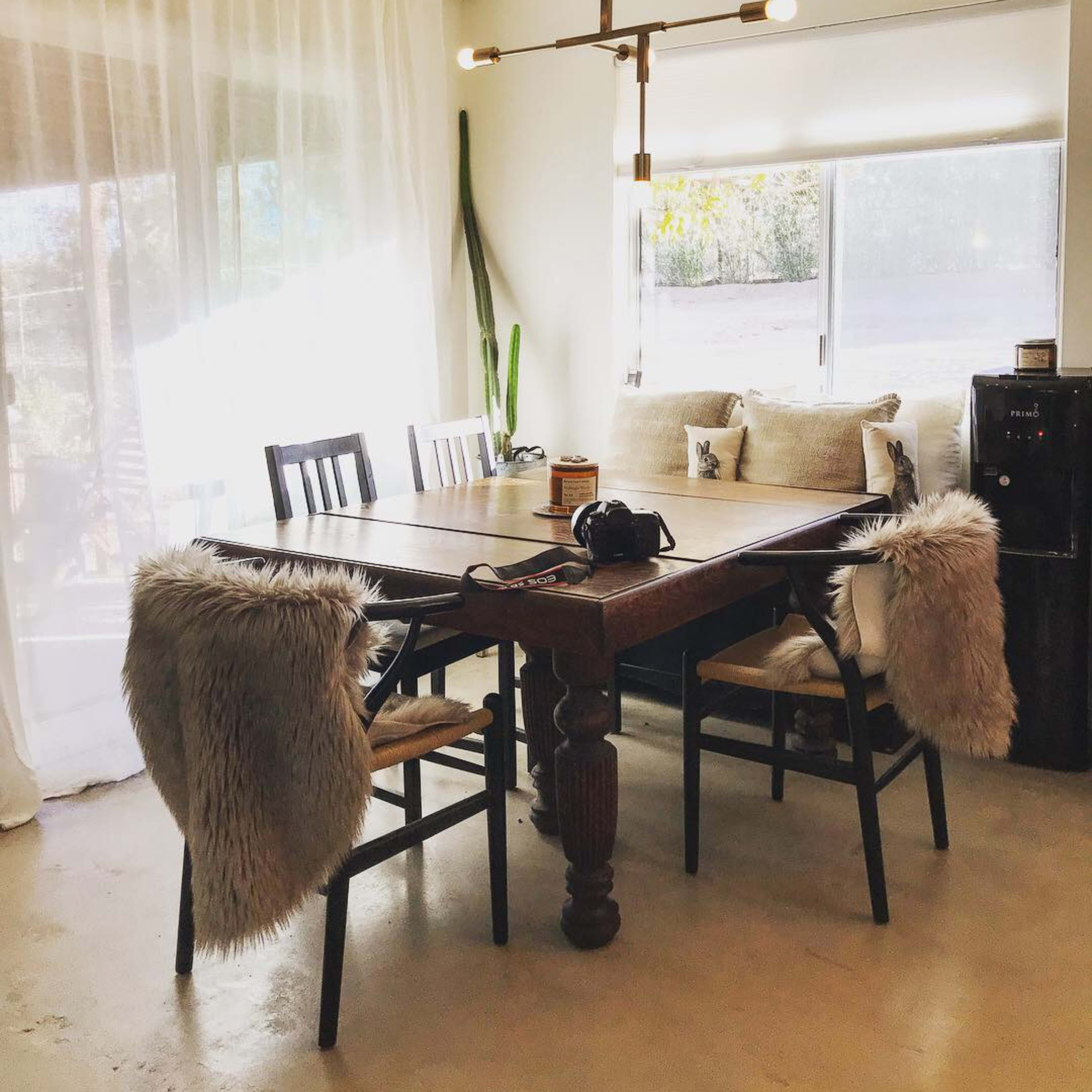 A wooden dining table with four chairs, two of which have fur throws, is set near a large window with sheer curtains and a cushioned bench.