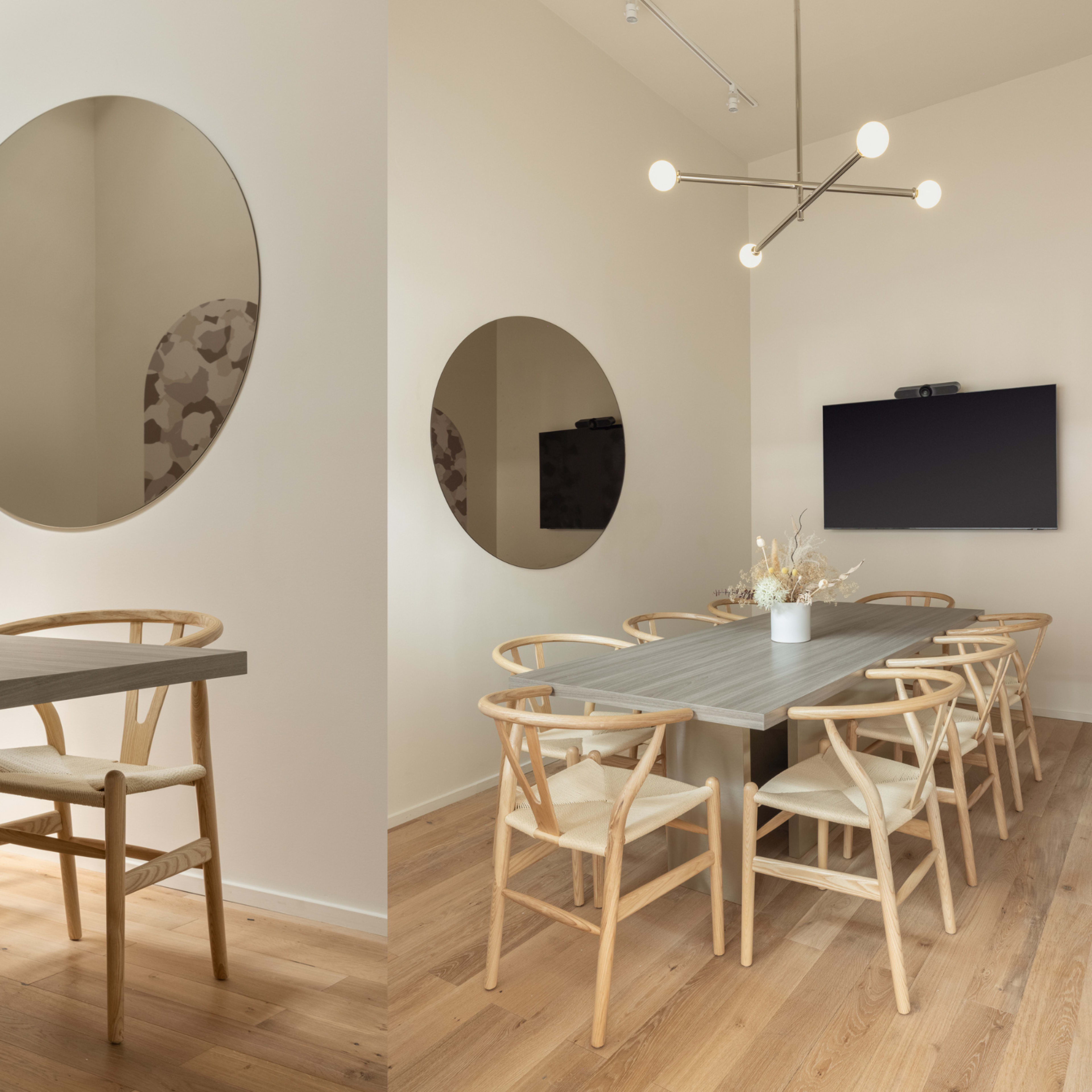 A modern dining area with a large oval mirror, a simple wooden table, and several light-colored chairs arranged around it, alongside a wall-mounted television.