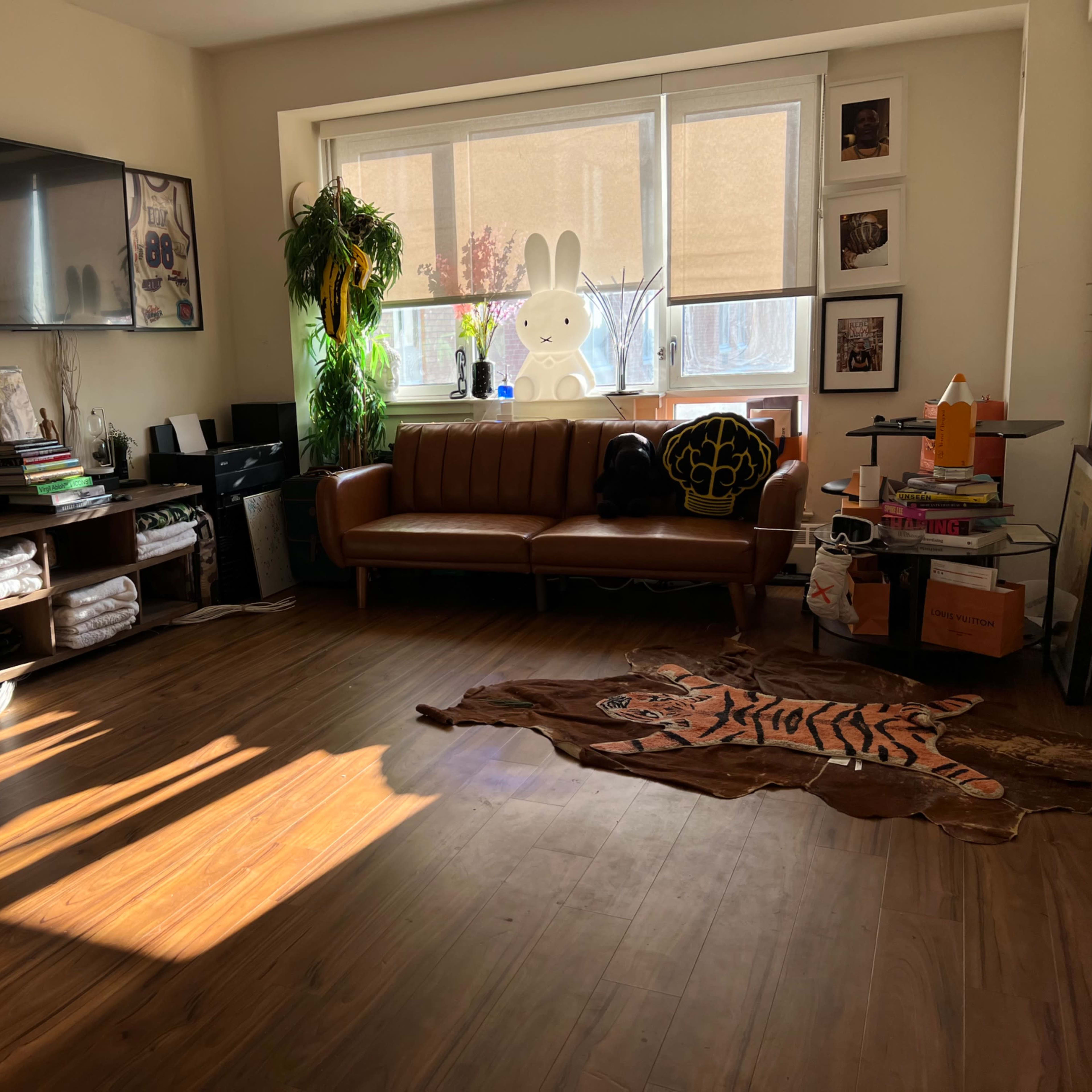 A well-lit living room with a brown leather couch, a television mounted on the wall, a large white rabbit statue, and a tiger-patterned rug on the wooden floor.
