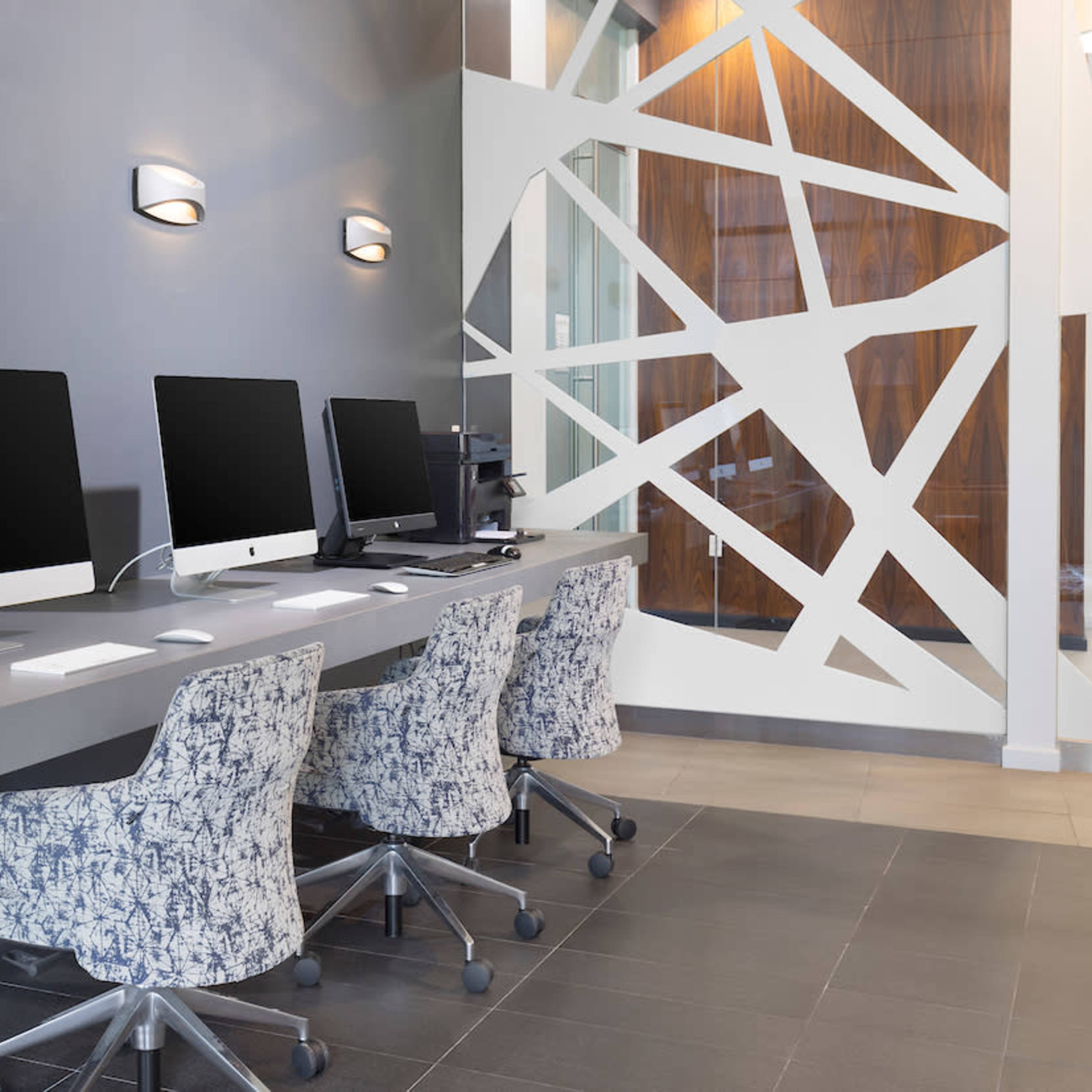 Three desktop computers are positioned on a sleek, light-colored desk, accompanied by patterned chairs, against a modern gray wall.