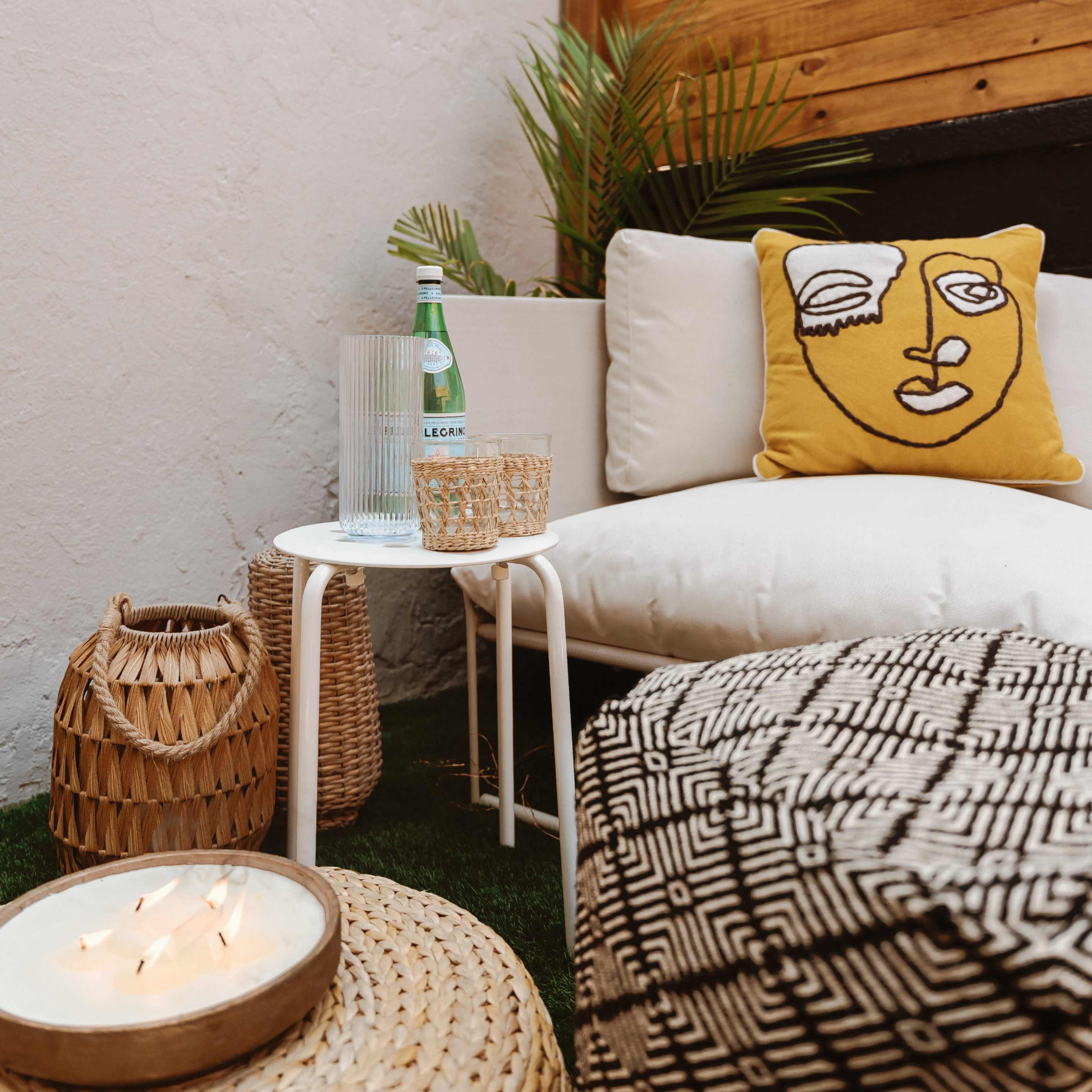 A cozy outdoor seating area features a white couch with a decorative yellow pillow, a small table with a water bottle, and various woven accessories.