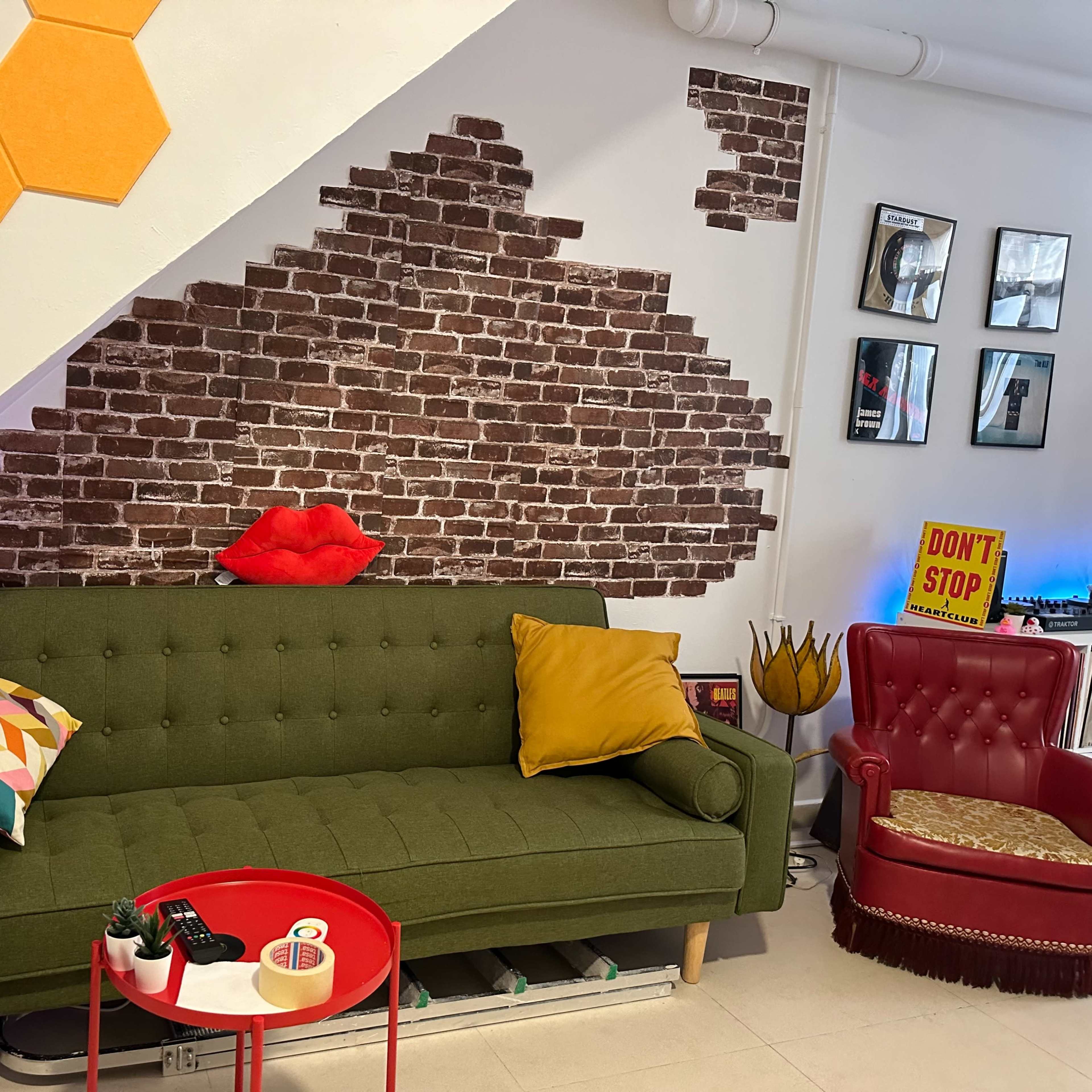 A green sofa with decorative cushions sits in front of a brick-patterned wall, accompanied by a red accent table and a vintage red chair.
