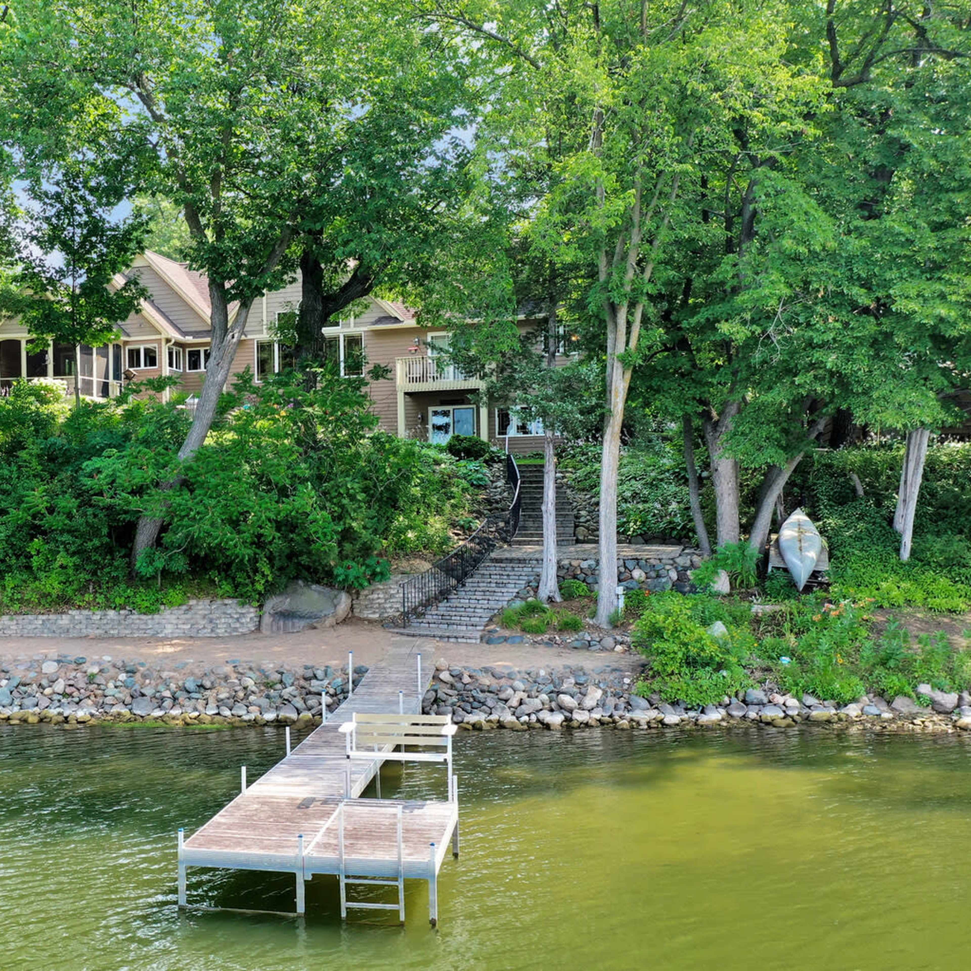A wooden dock extends into a calm body of water, with a house and lush greenery visible along the shoreline.