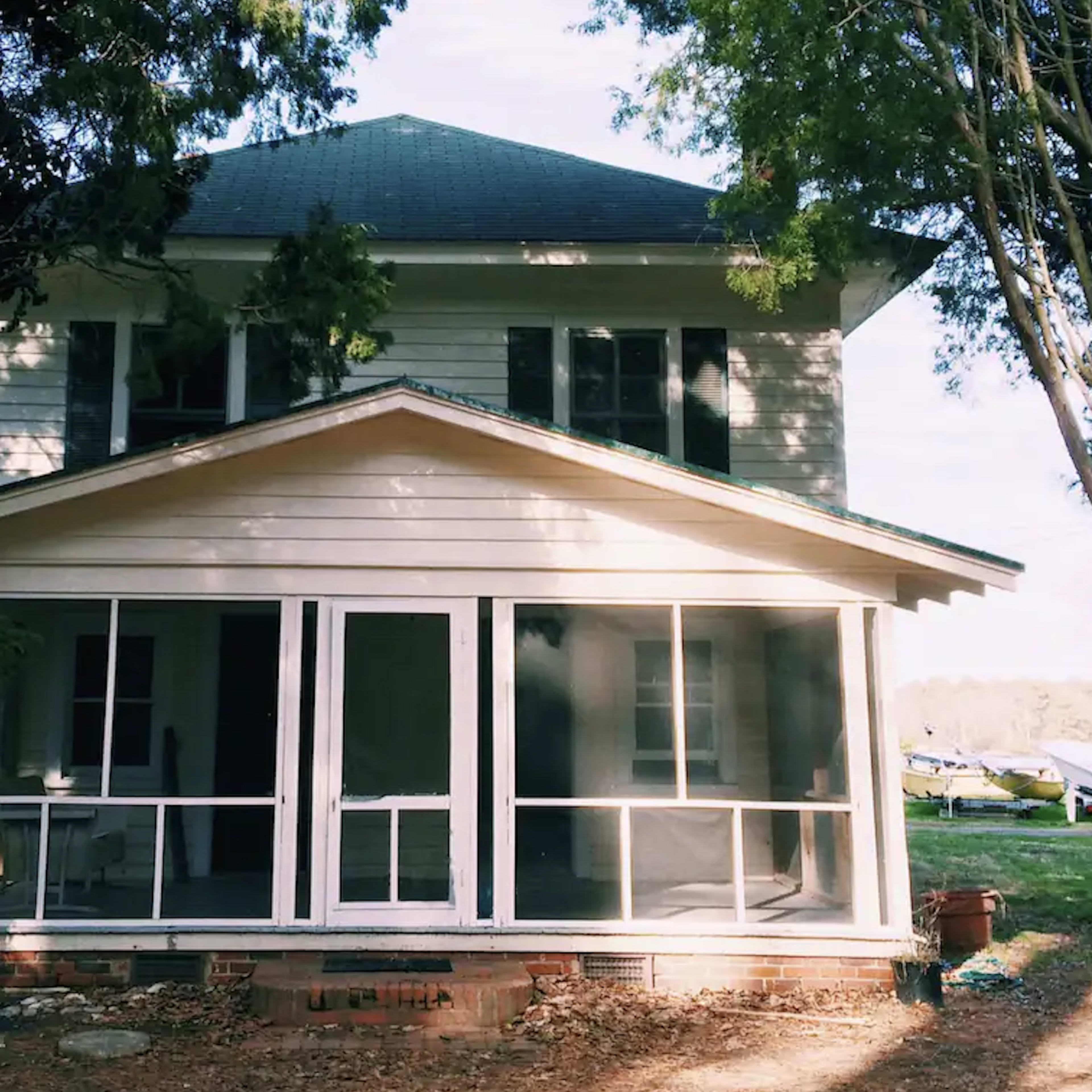 A two-story wooden house with a front porch featuring screens, surrounded by trees.