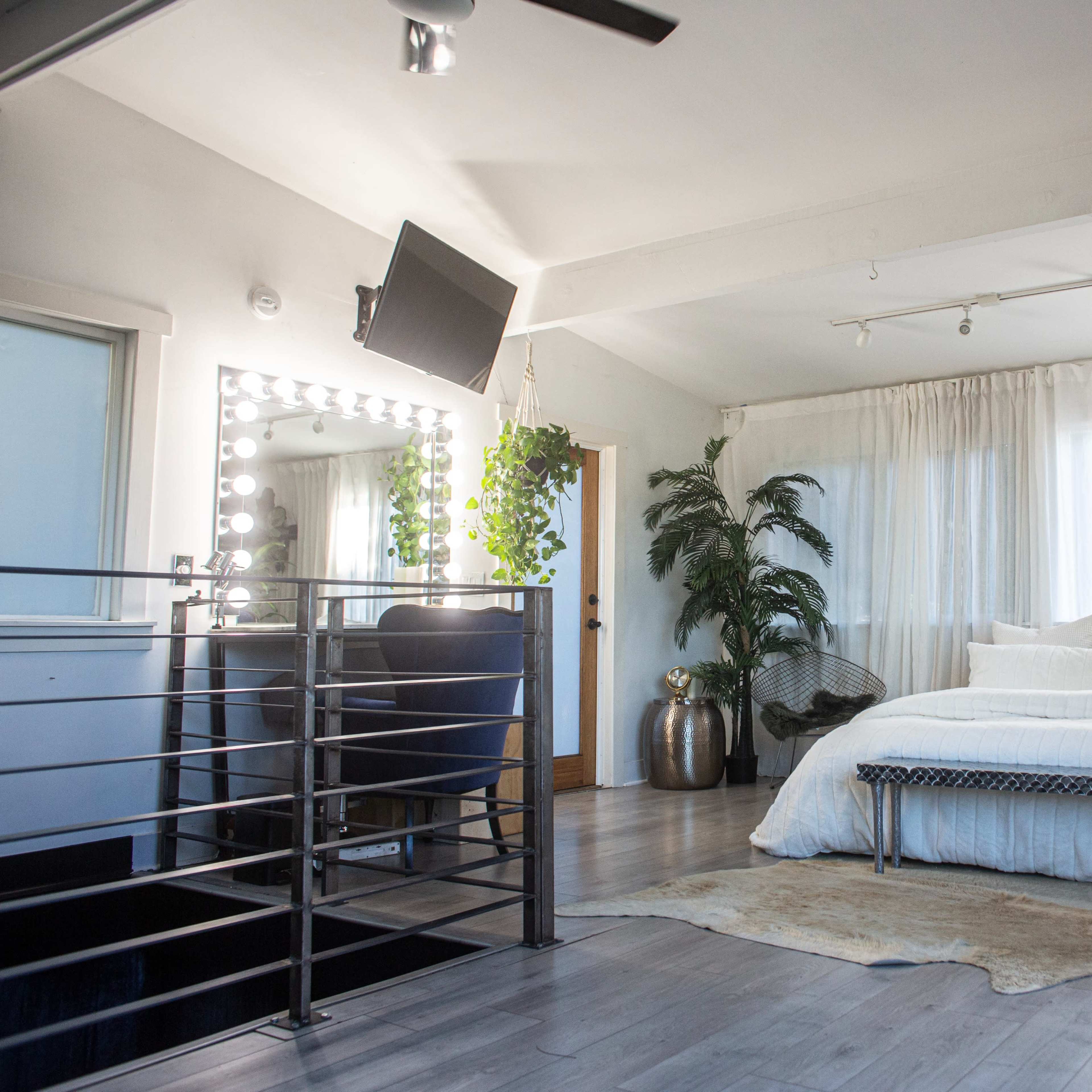 A spacious bedroom featuring a large bed with white linens, a mirror-lined vanity area, and plants placed throughout the room.