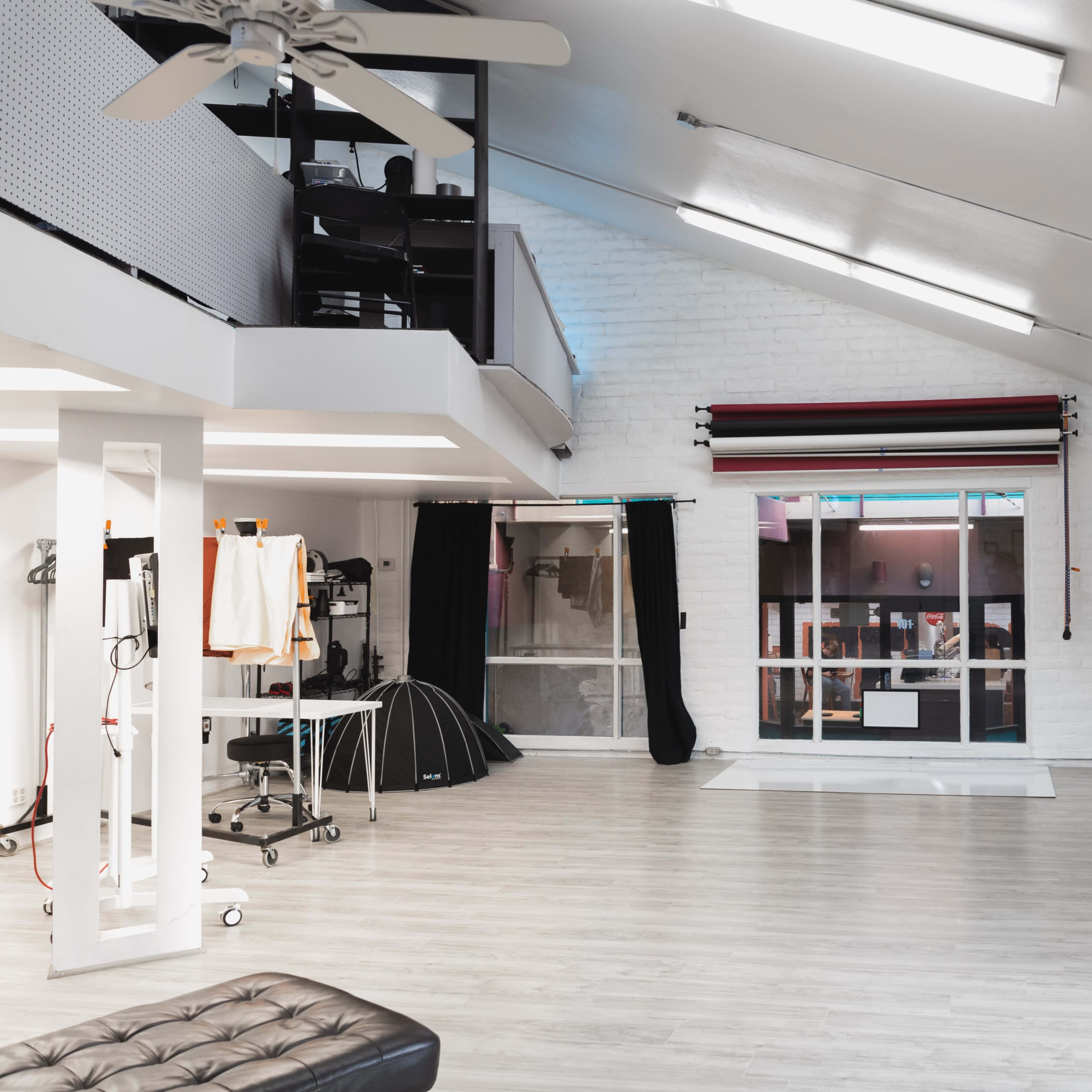 A spacious, well-lit studio with white walls, wooden flooring, and large windows, featuring various furniture and equipment along the sides.