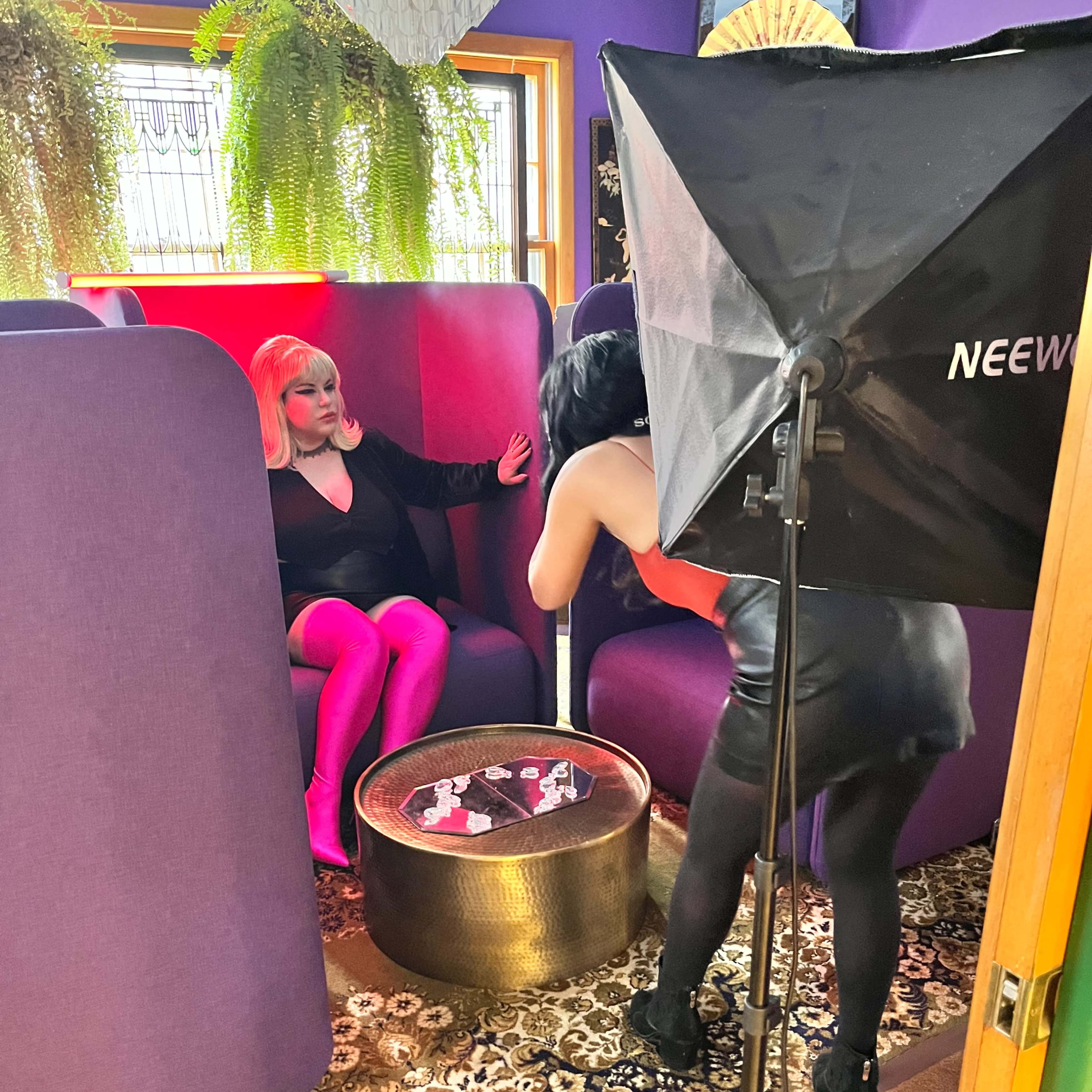A person with pink hair sits in a purple booth while another person with dark hair takes a photograph using a studio light setup.