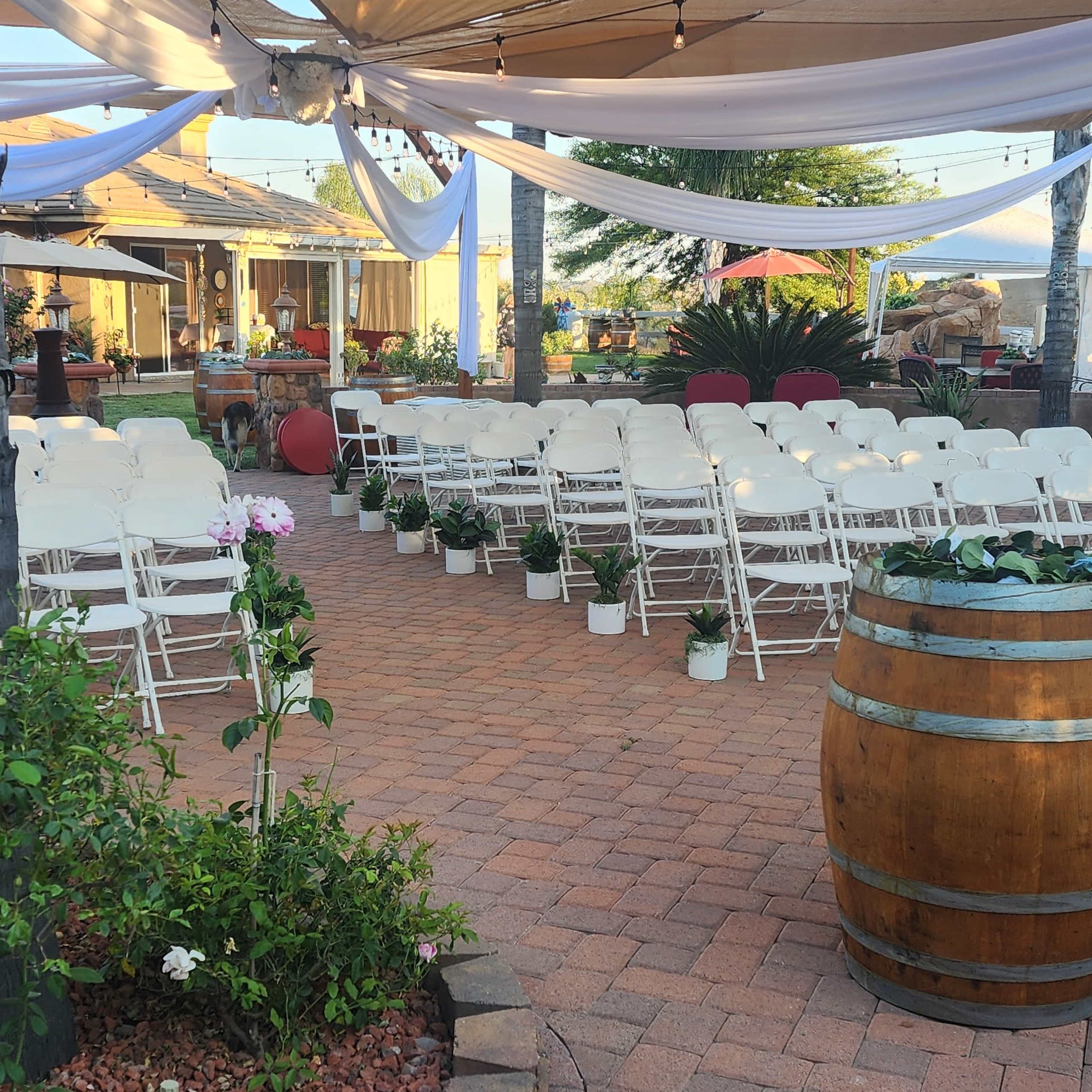 A prepared outdoor event space with rows of white chairs arranged on a brick patio, surrounded by potted plants and decorative elements.
