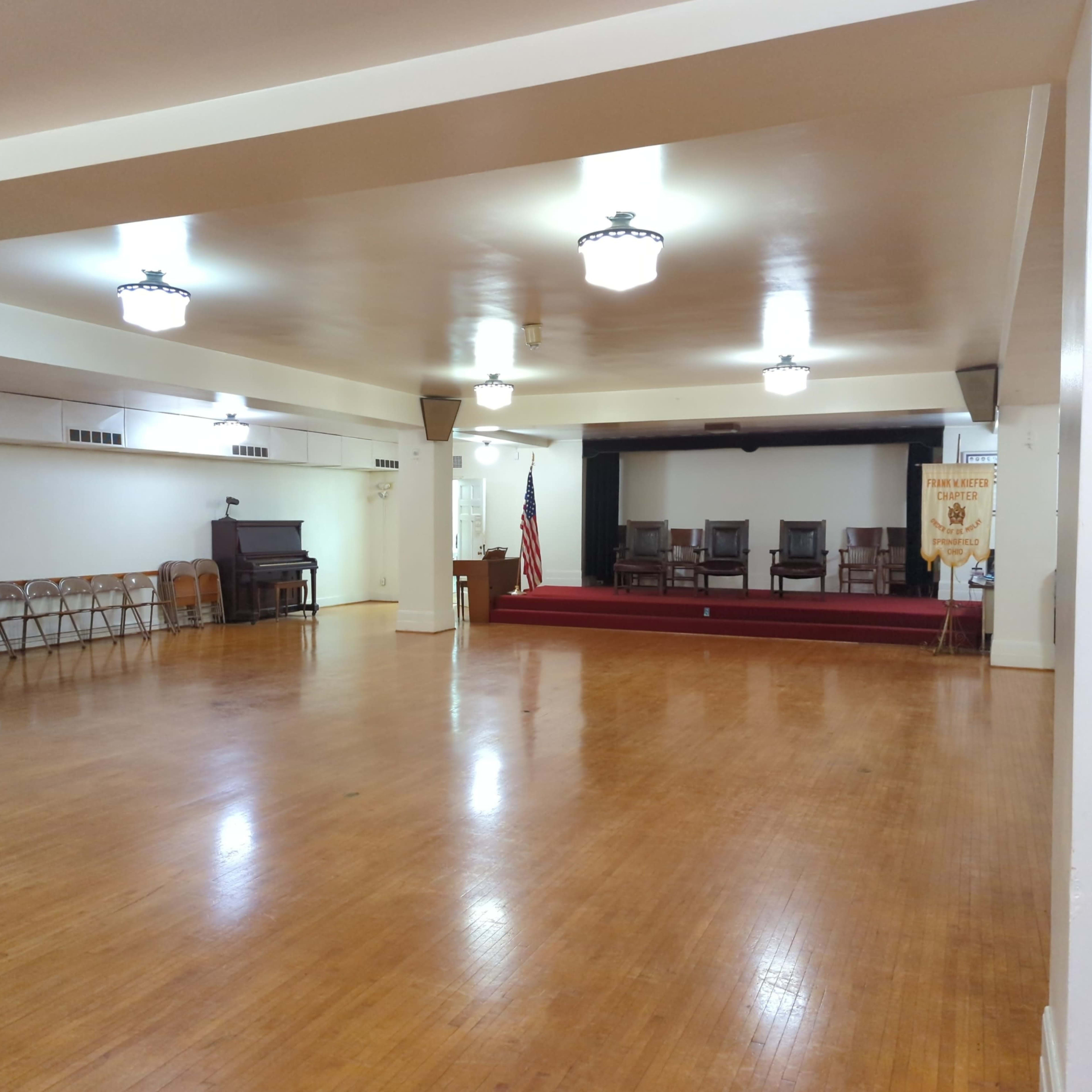 A spacious hall with wooden floors, a set of empty chairs lined against one wall, a podium at the front, an American flag, and a small stage with three chairs on a raised platform.