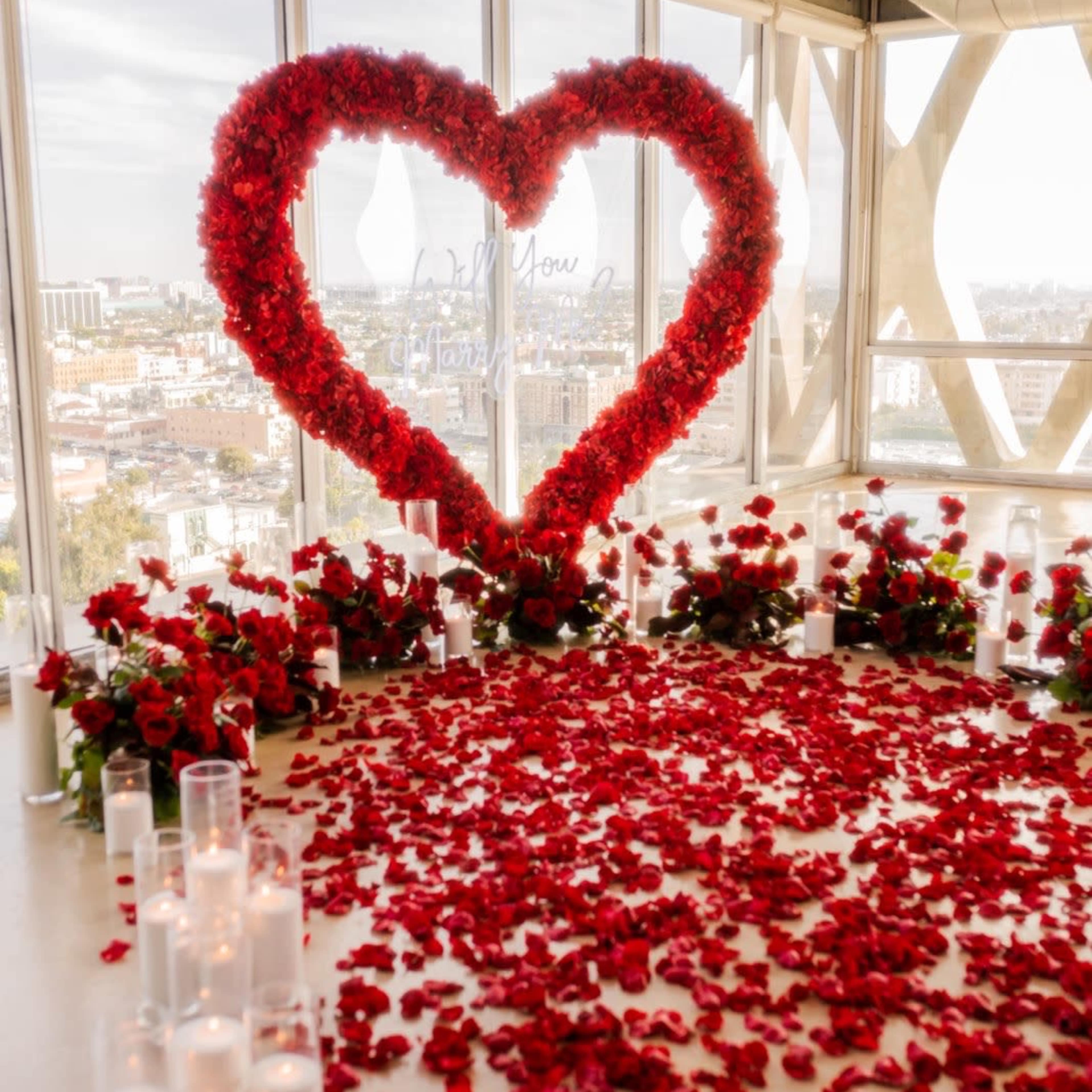 The scene features a large heart-shaped arrangement made of red flowers, surrounded by a carpet of rose petals and illuminated by candles, set against a backdrop of a city skyline through large windows.