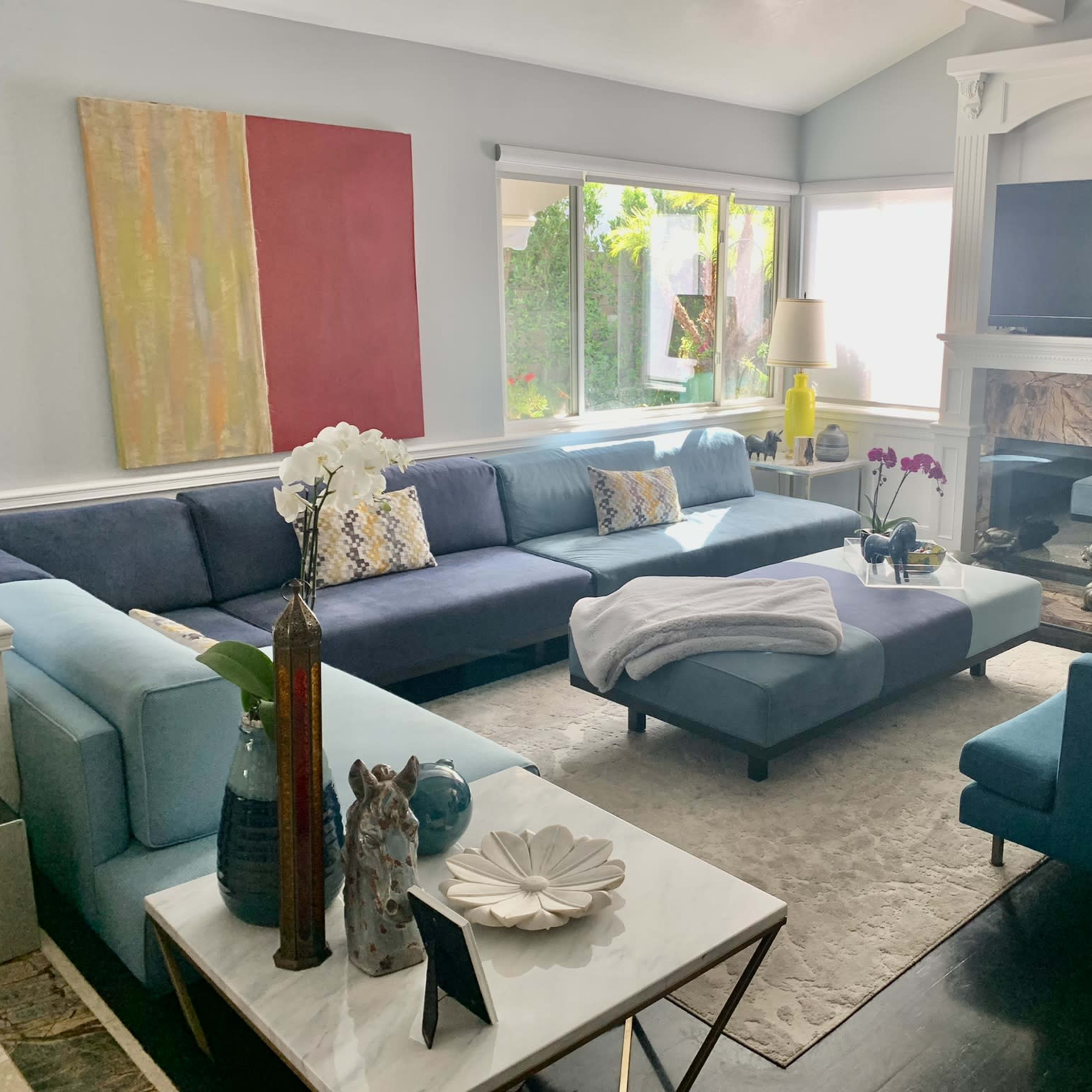 The living room features a large blue sectional sofa, a white coffee table, and a stone fireplace, with windows providing natural light and plants visible outside.