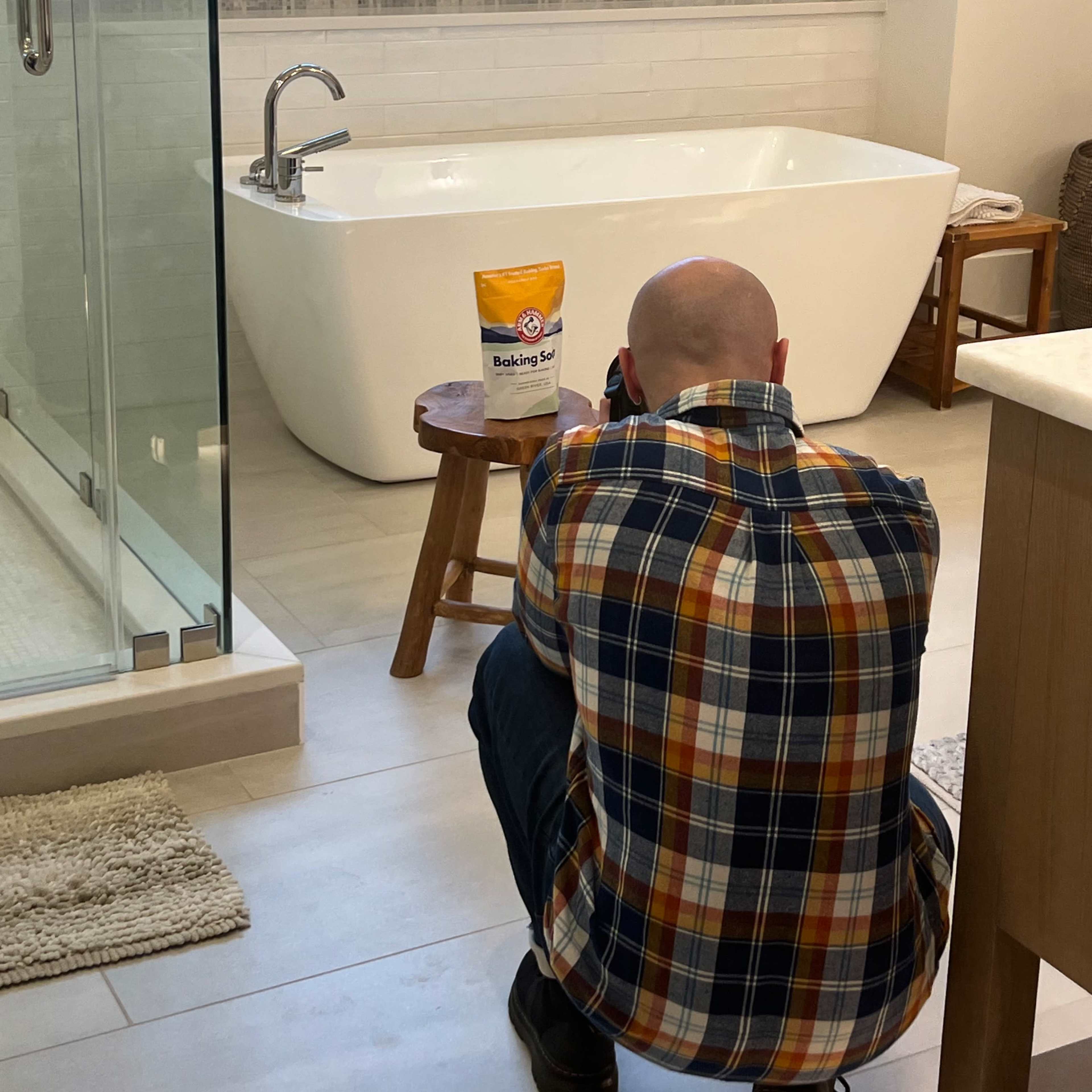 A person in a plaid shirt is crouched down taking a photograph of a container of baking soda placed on a small stool in a bathroom.