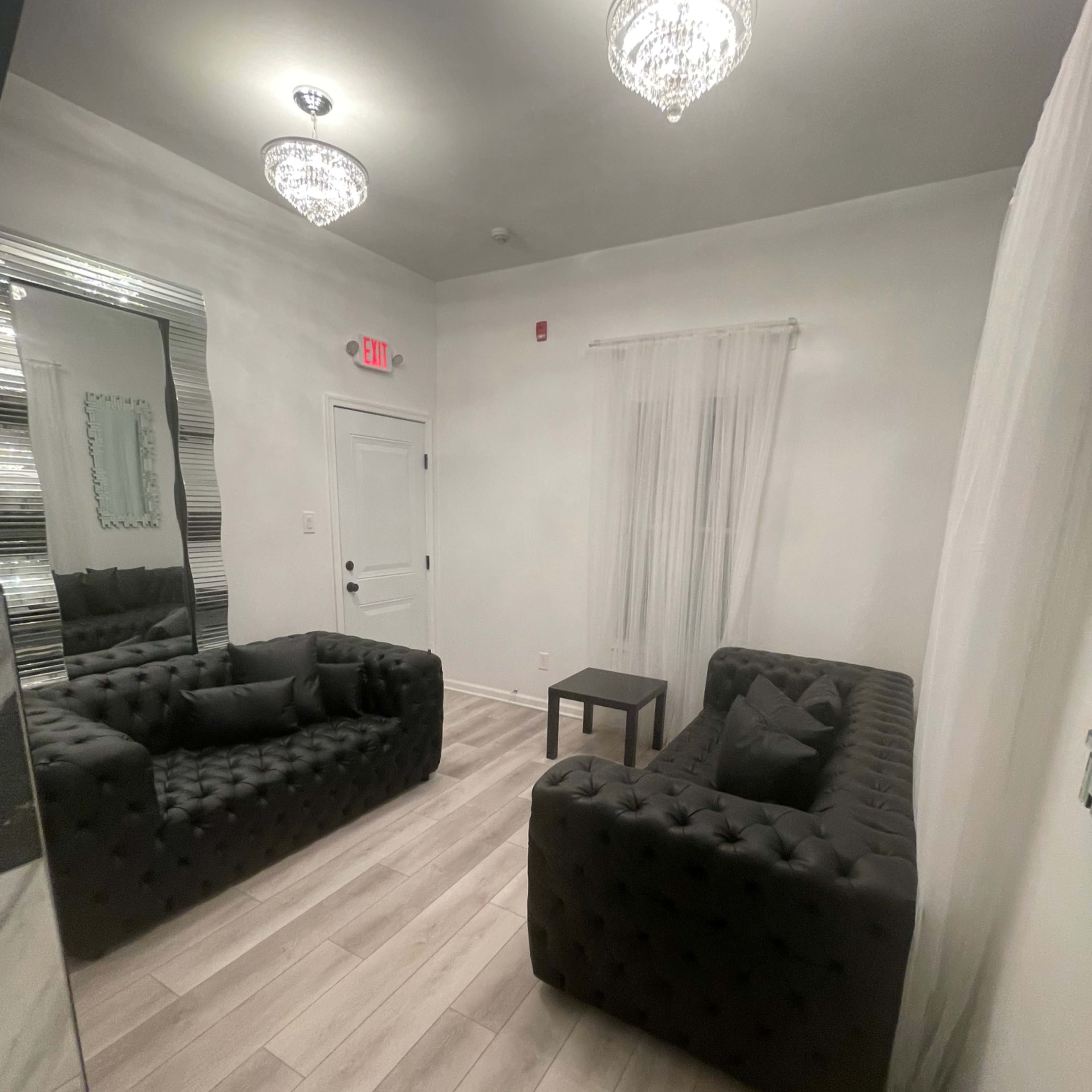 A minimalistic room with two black tufted sofas facing a small table, accented by two crystal chandeliers and a mirror on one wall.