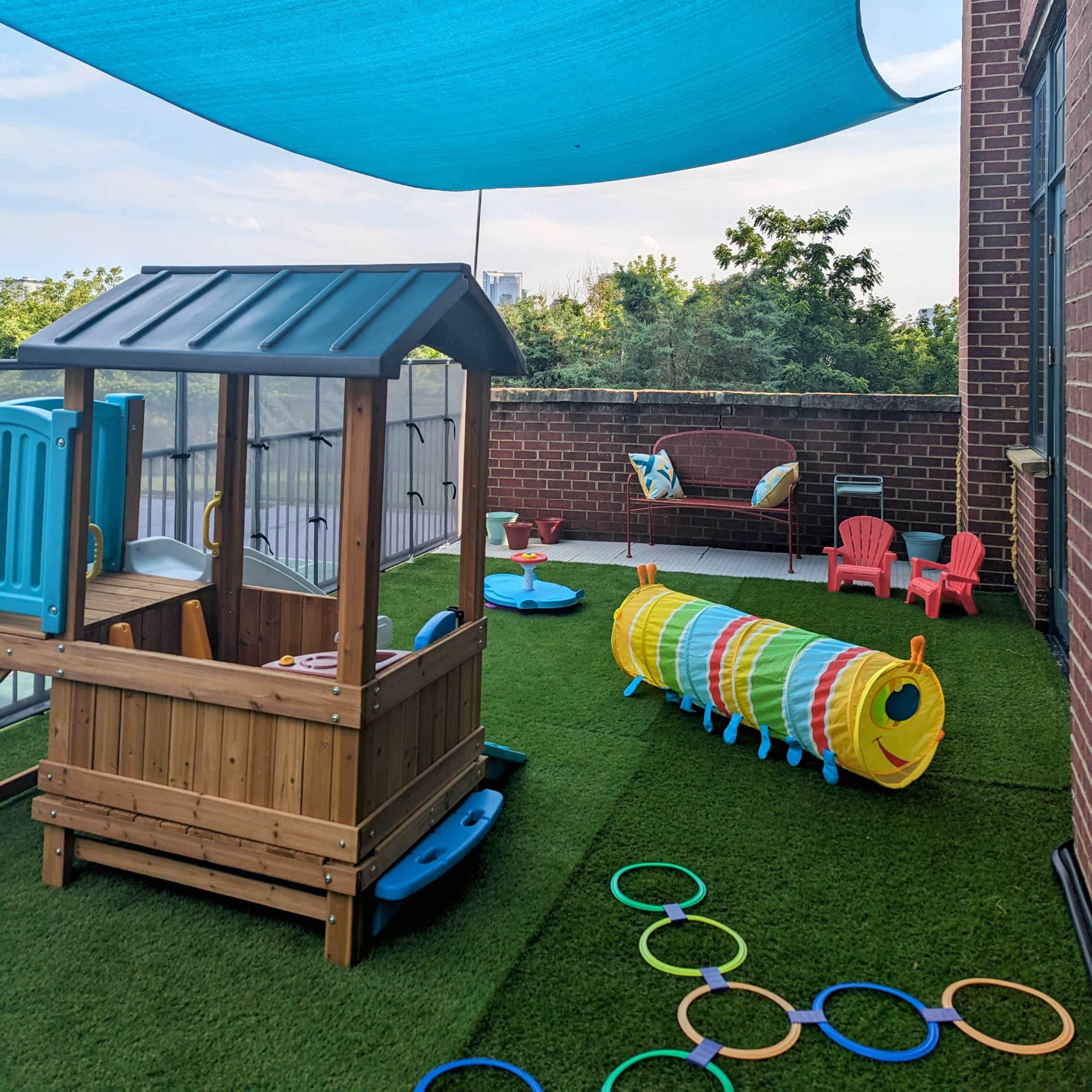 A play area on a balcony featuring a wooden playhouse, colorful tunnel, and various toys under a blue shade sail.