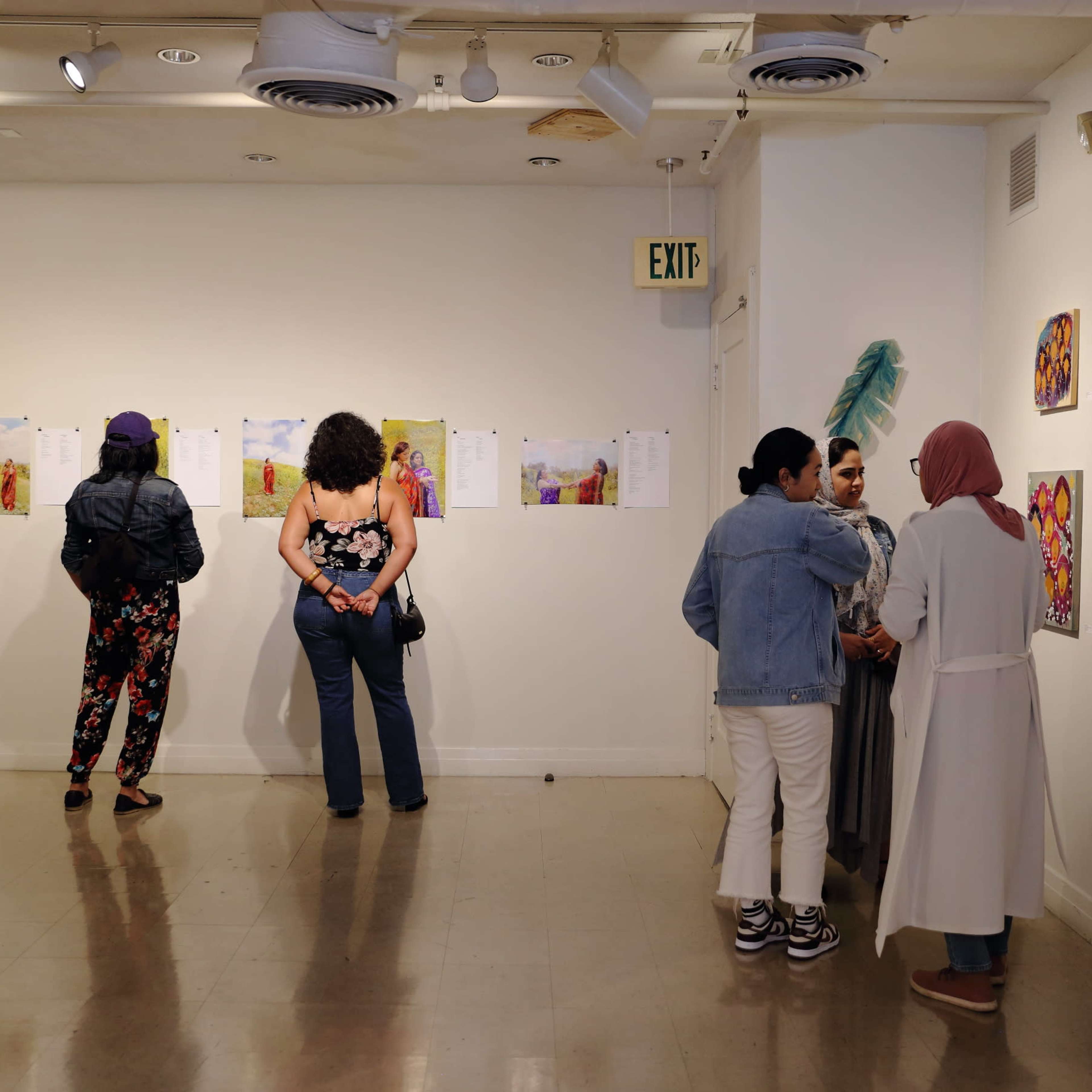 A group of people stands in an art gallery, observing artworks displayed on the wall while others engage in conversation.