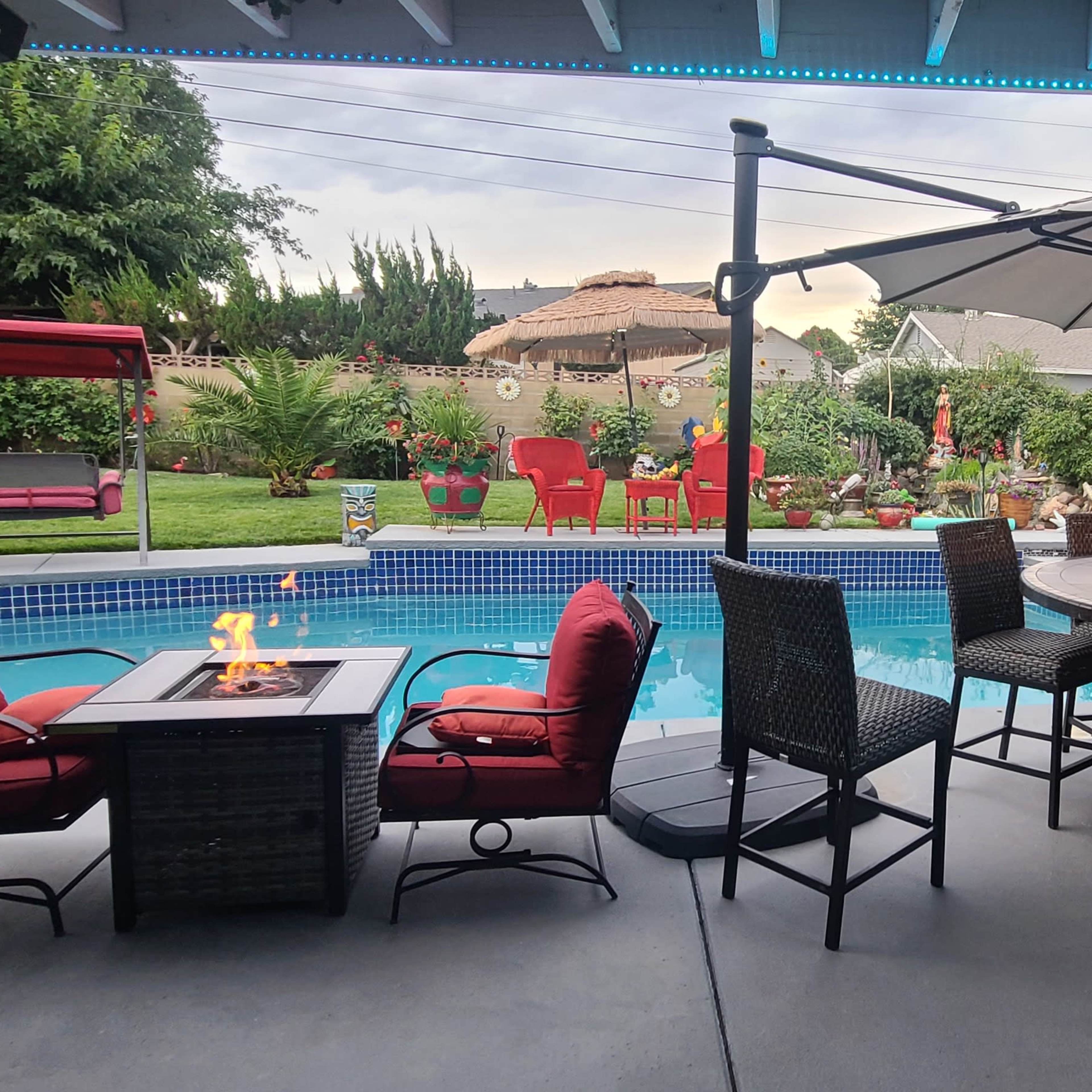 A backyard patio setting with a swimming pool, a large umbrella over a table with flames, and several red chairs and a swing in the garden area.