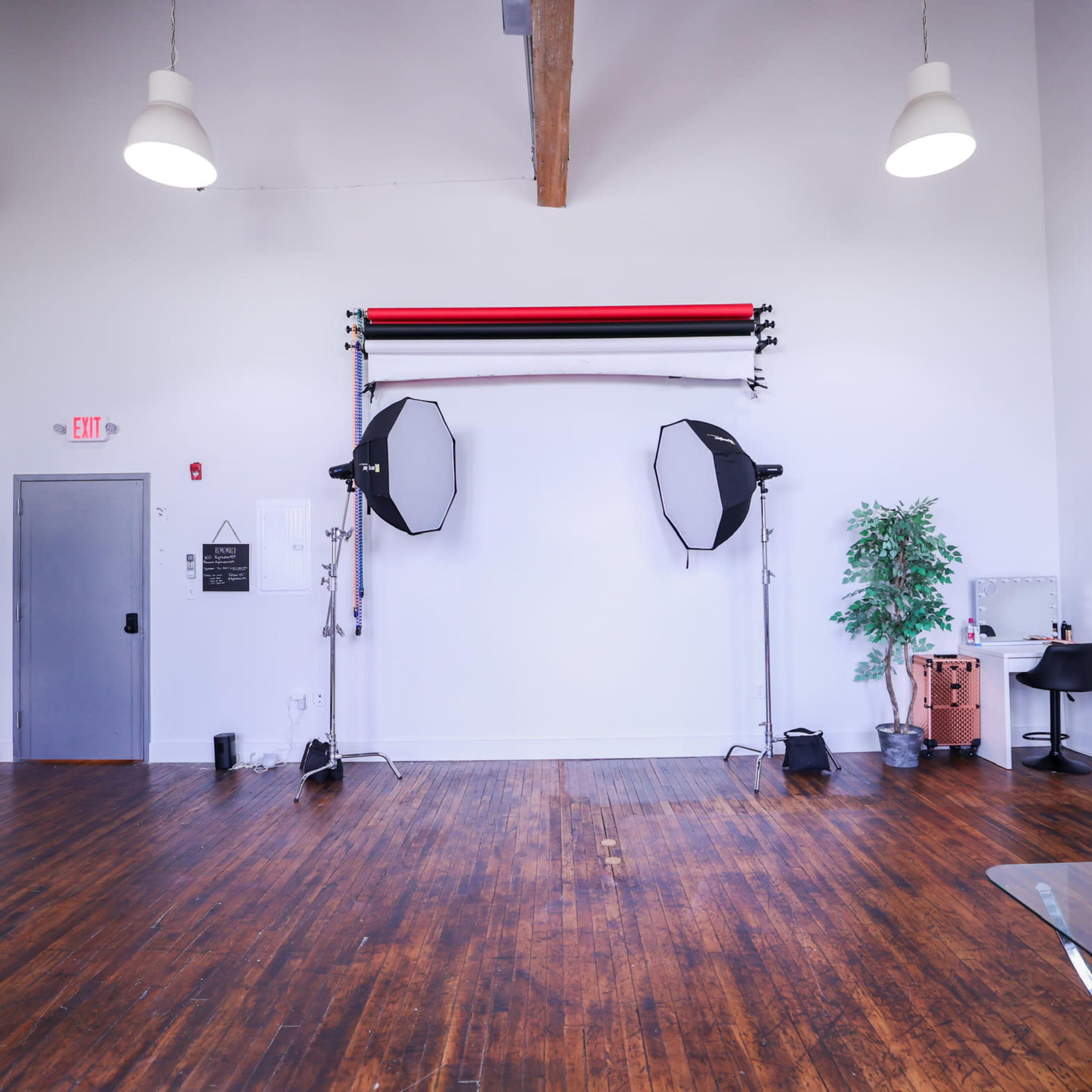 A spacious photography studio featuring backdrop stands with lights, a green plant, a vanity area, and a red couch on wooden flooring.
