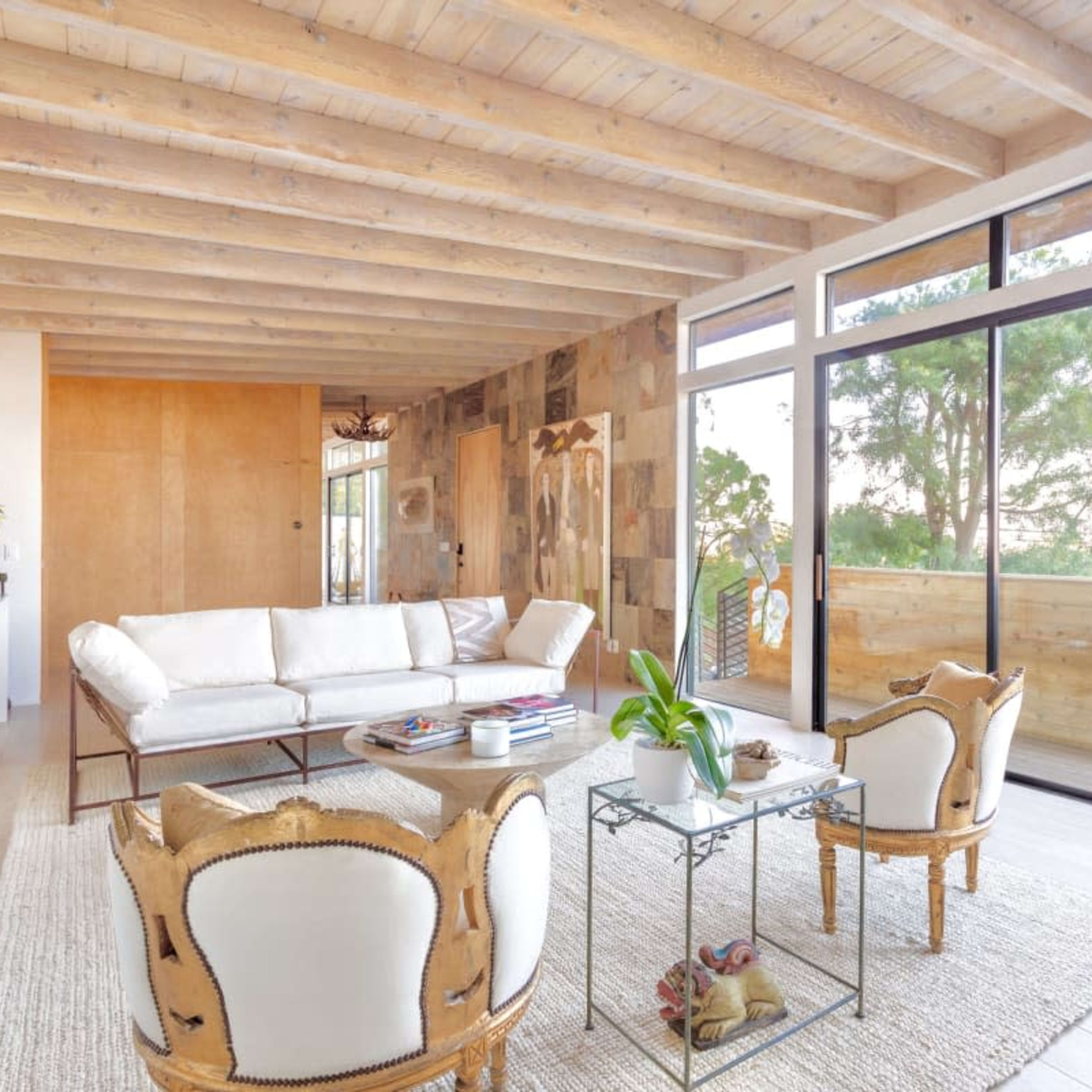 A modern living room with a white sofa, two patterned chairs, a coffee table, and large windows facing a wooden deck and outdoor greenery.