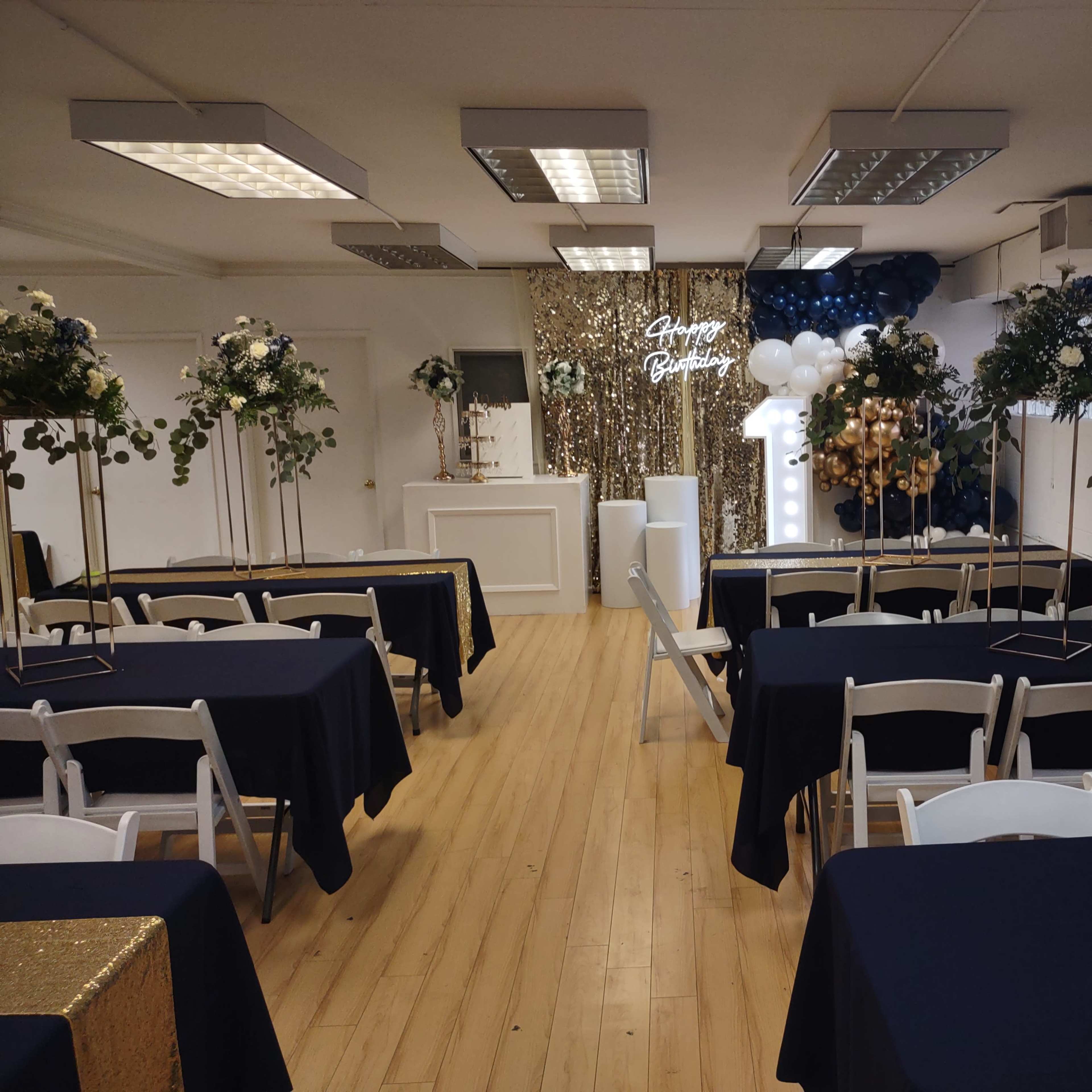 A decorated event space features tables covered with navy cloths, surrounded by greenery, with a backdrop of balloons and a sparkling golden curtain.