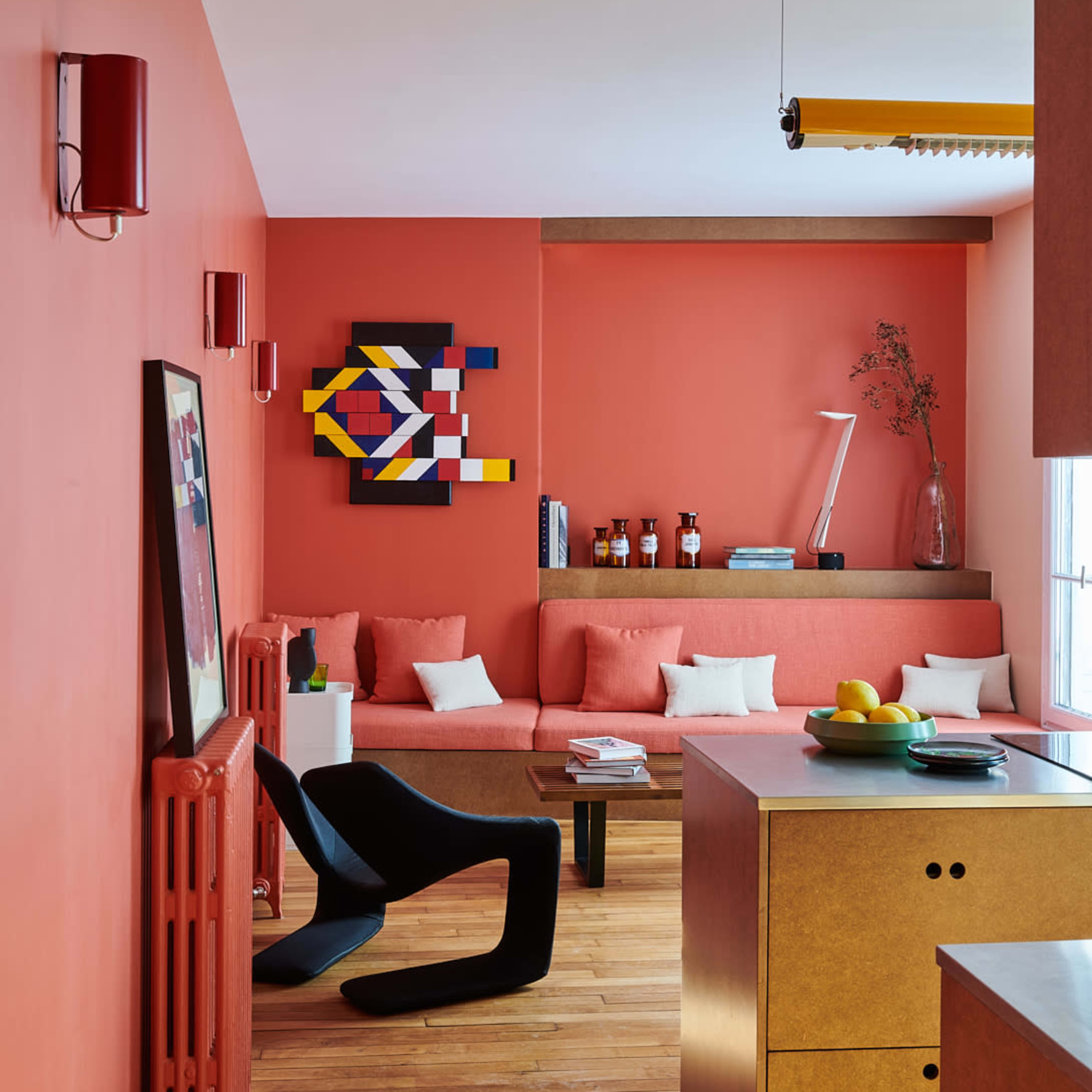A modern living space with a coral-colored wall, a sectional sofa, and a wooden kitchen island, featuring geometric artwork and decor items.