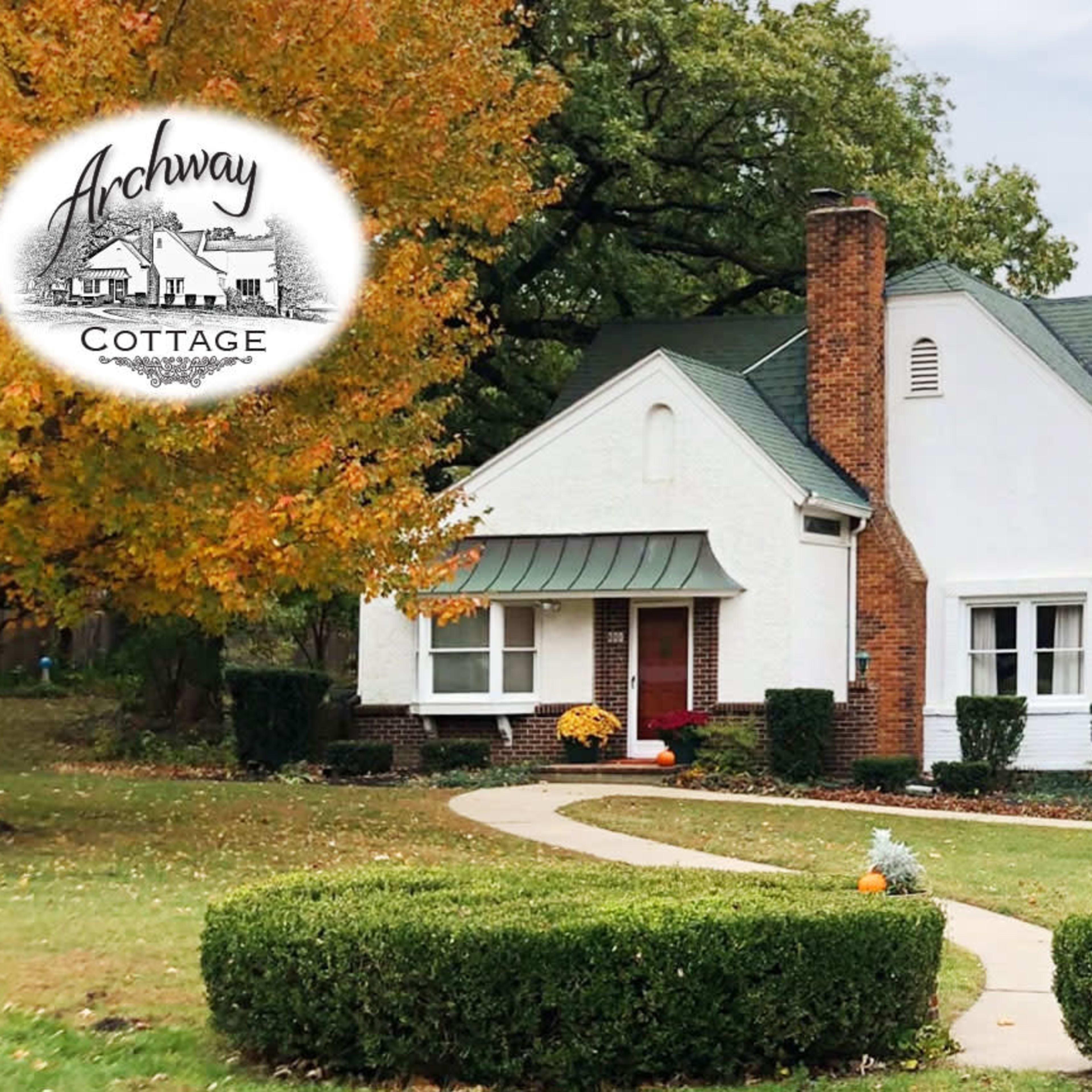A white cottage with a green roof is surrounded by landscaping and fall-colored trees.