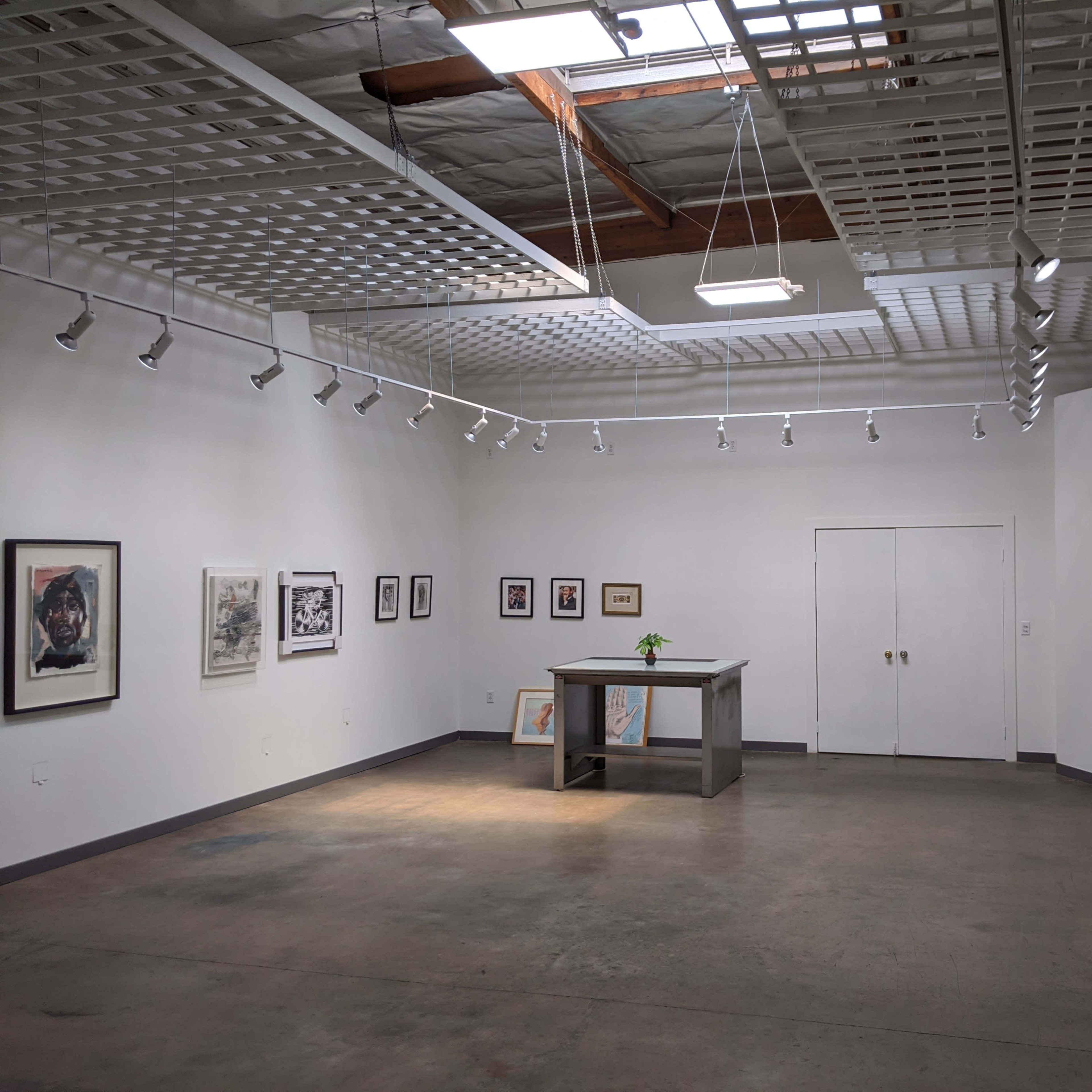 An art gallery features white walls, a central gray display table with a small plant, and framed artworks along the sides.