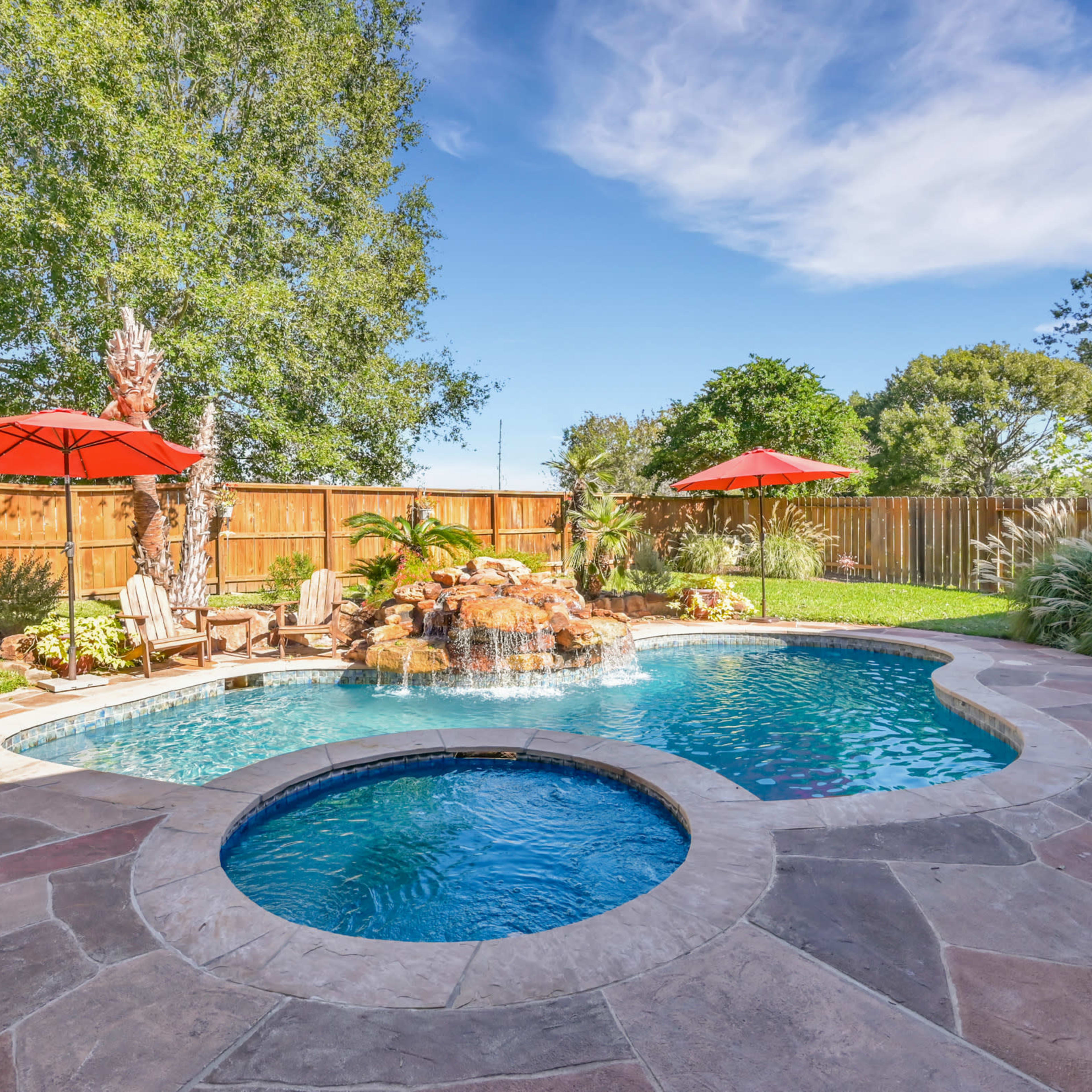 A backyard with a swimming pool featuring a rock waterfall, surrounded by chairs, umbrellas, and landscaped greenery.