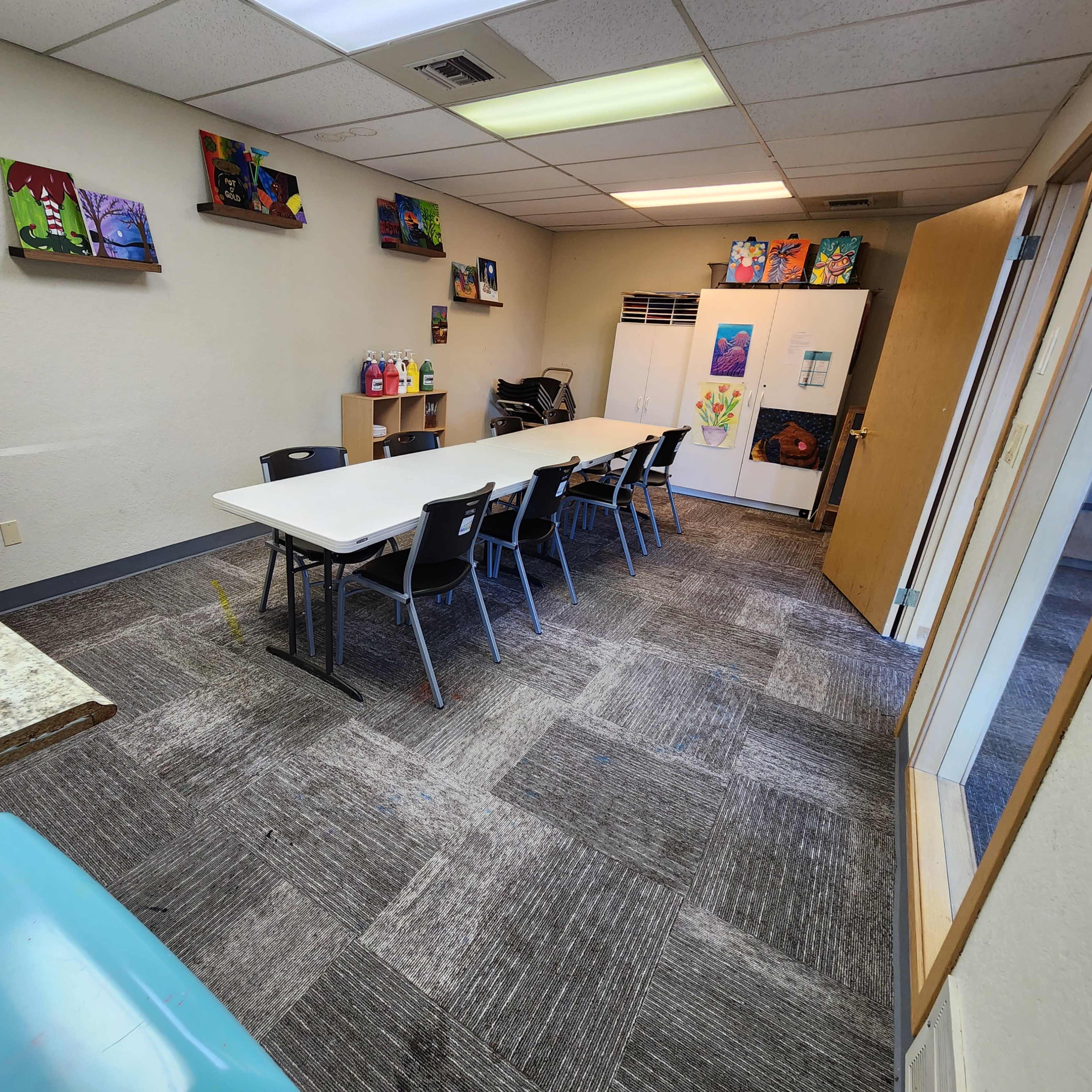 A long table with chairs is situated in a room adorned with colorful wall art, featuring a kitchenette and a refrigerator in the background.
